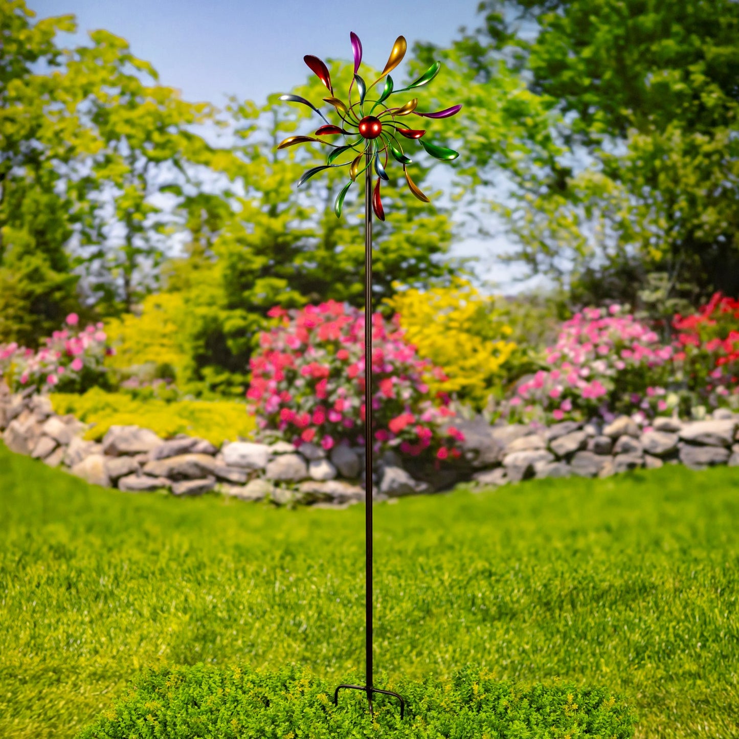 A colorful metal garden wind spinner on a black stake, set in a lush green lawn with a blurred background of pink flowers and trees.