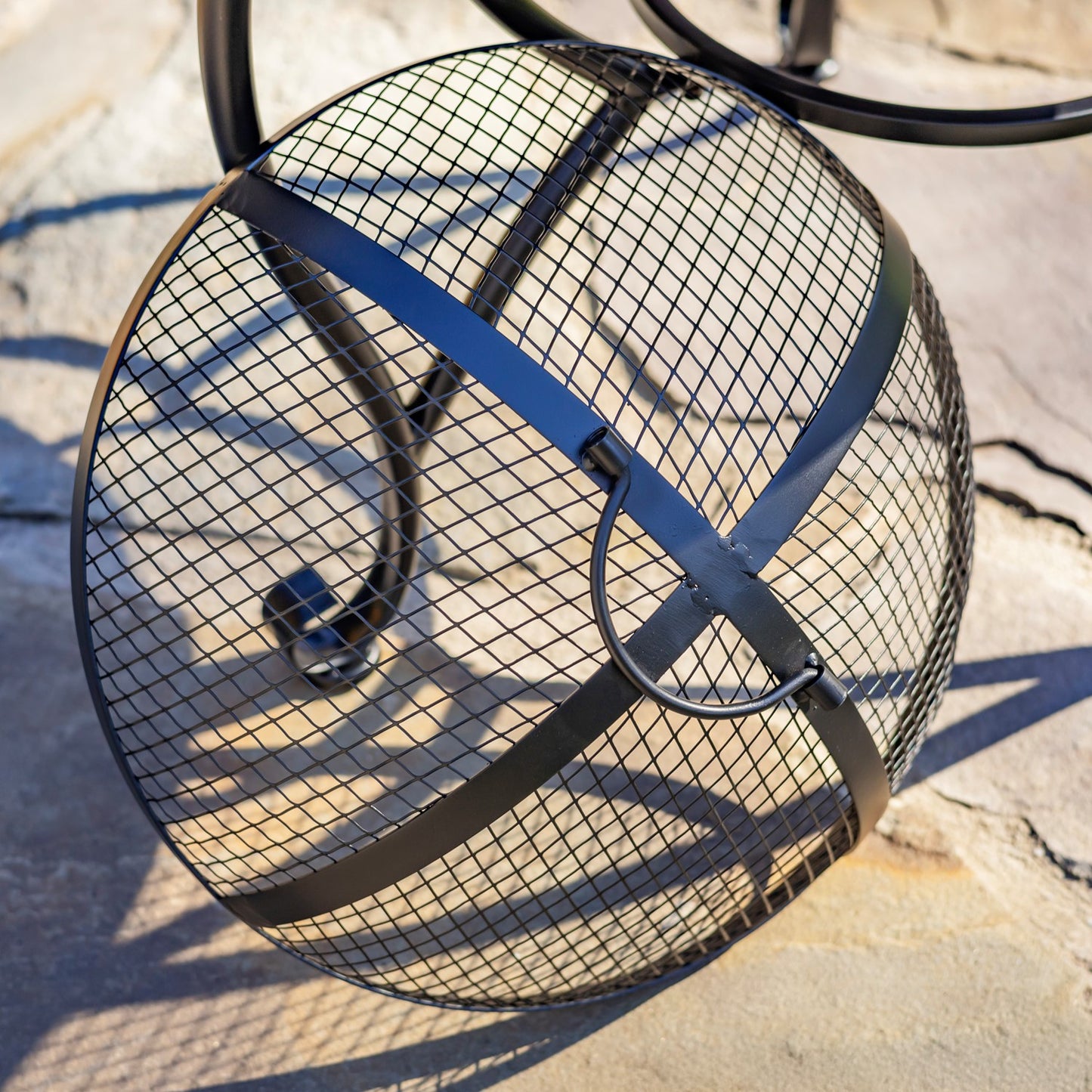 A black metal fire pit basket with a wire mesh sphere design and curved support bars, photographed outdoors on a stone surface with strong sunlight and shadows.