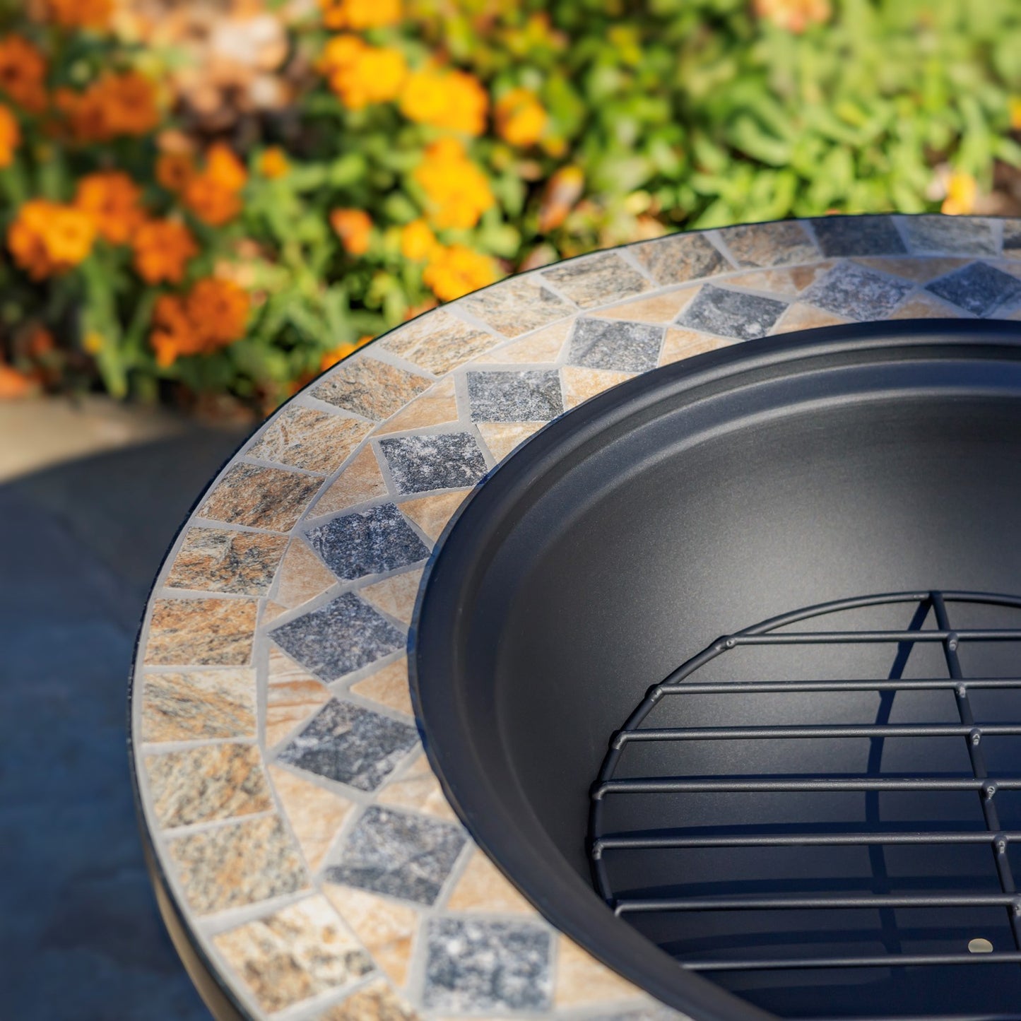 Close-up of a round outdoor fire pit table with a black interior bowl and a mosaic tile rim in beige, gray, and blue stones, with blurred flowers in the background.