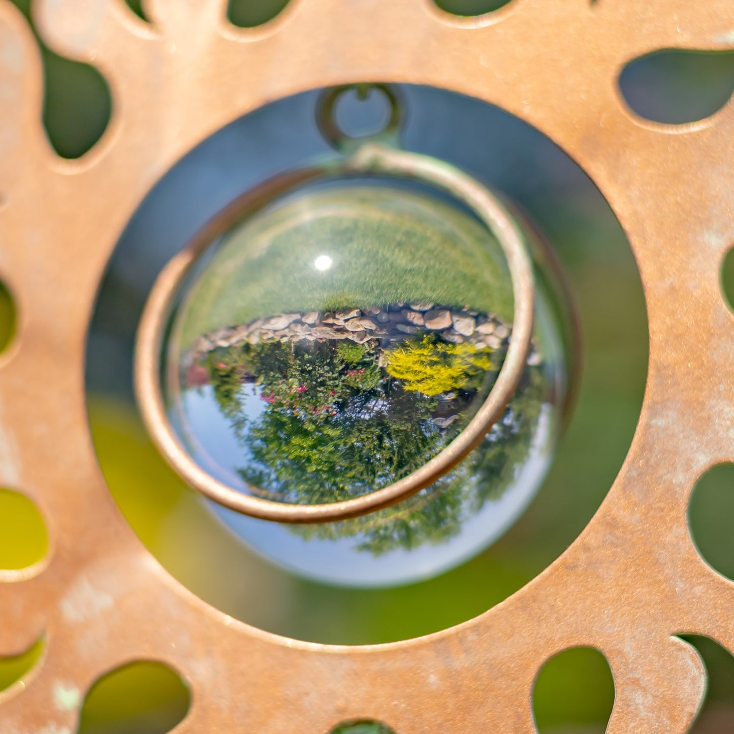 A close-up of a round glass ornament or bead with a small metal loop at the top, resting inside a circular cutout of a rusted metal ring. The glass reflects green foliage and sky.