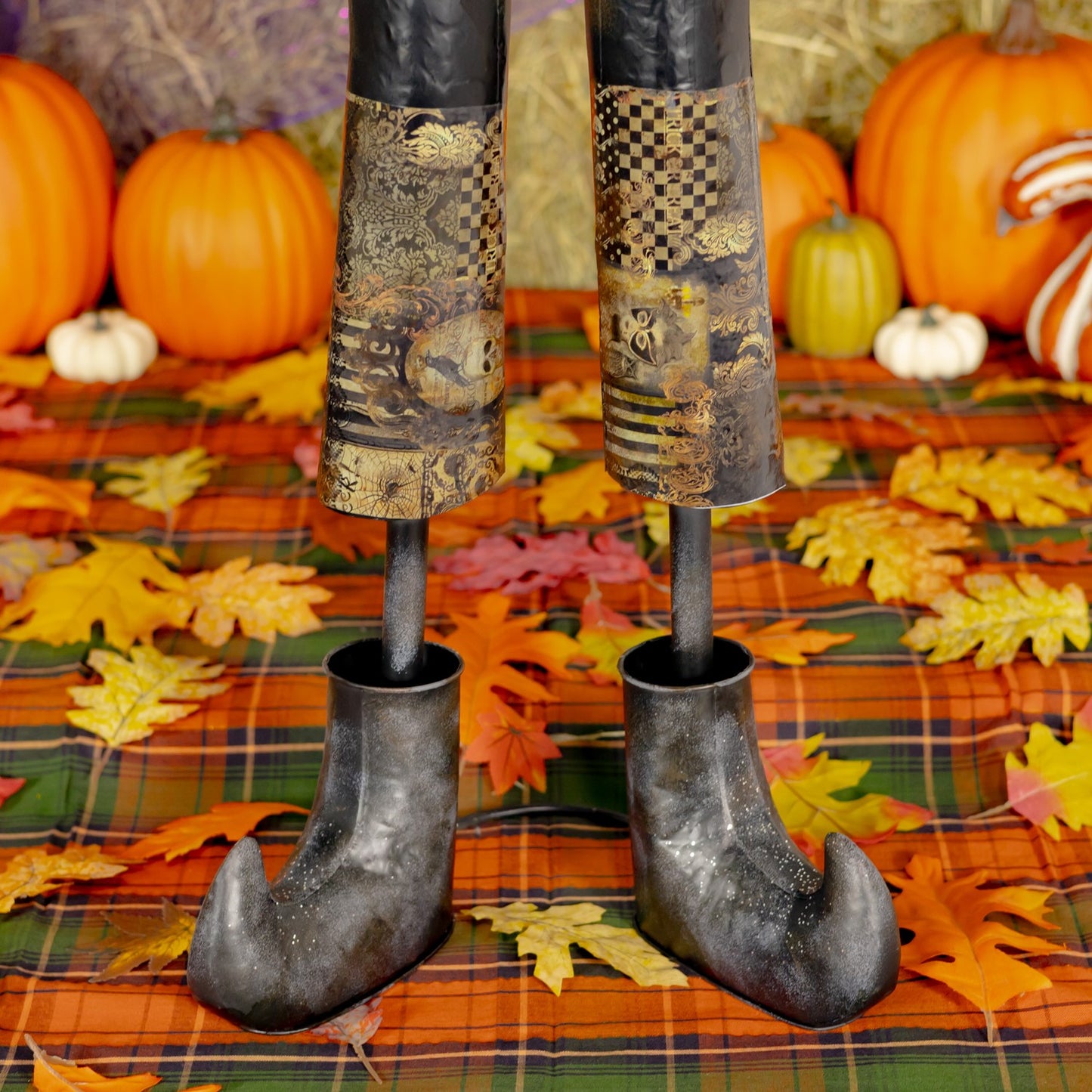 A Halloween decoration shaped like a witch's legs, with patterned black and gold stockings and pointed dark shoes, standing on a plaid blanket with autumn leaves, pumpkins, and hay bales in the background.