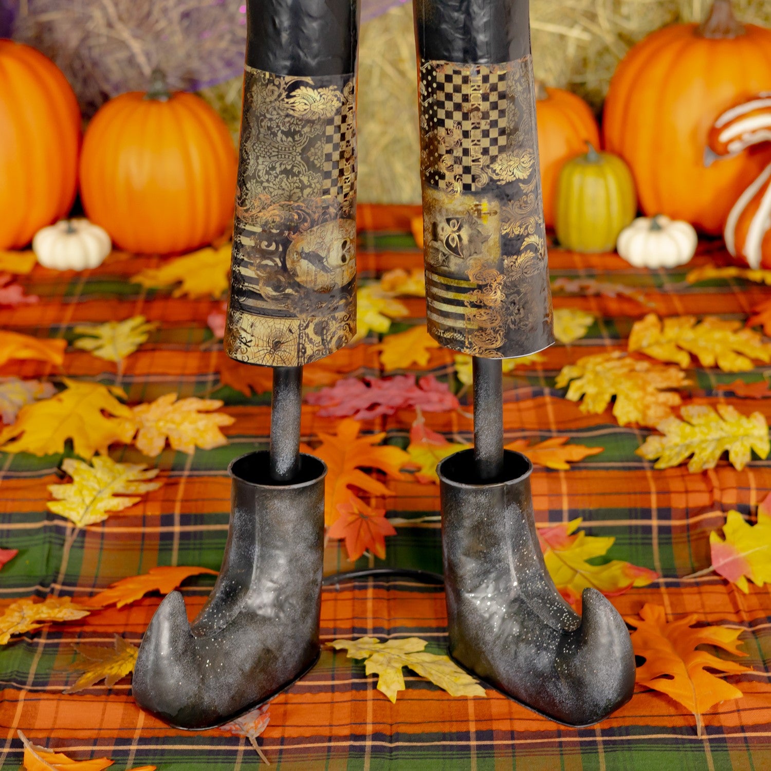 A Halloween decoration shaped like a witch's legs, with patterned black and gold stockings and pointed dark shoes, standing on a plaid blanket with autumn leaves, pumpkins, and hay bales in the background.