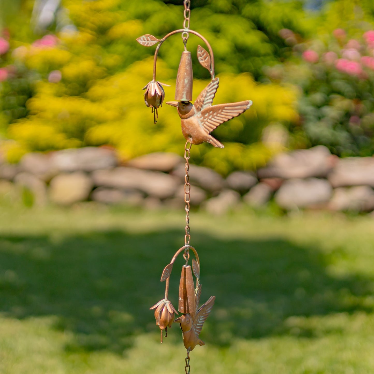 A copper garden wind chime featuring a hummingbird and seed pods, hanging outdoors against a blurred green garden background.