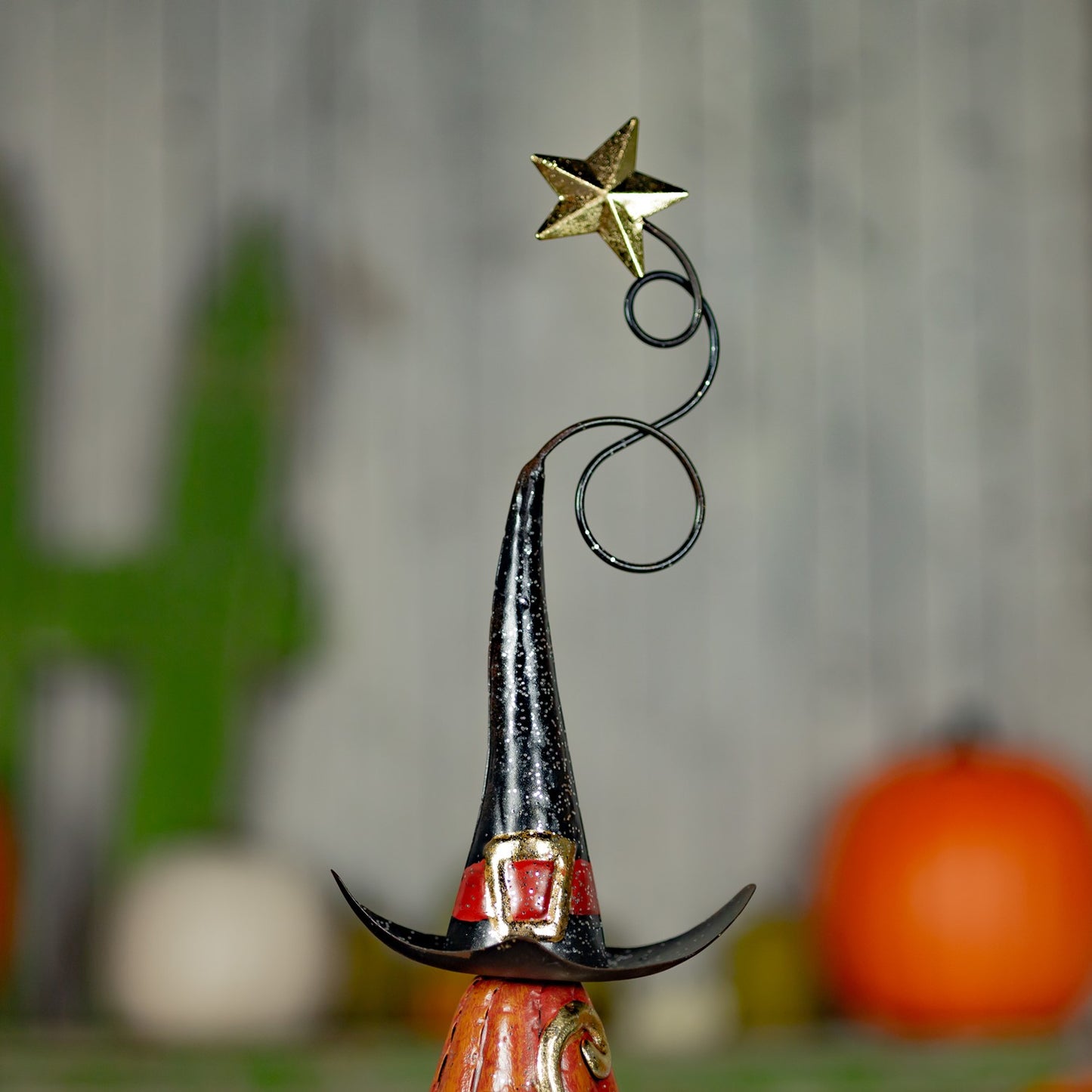 A decorative metal witch hat ornament with a curved black wire stem topped by a gold star, shown against a softly blurred autumn background with pumpkins.