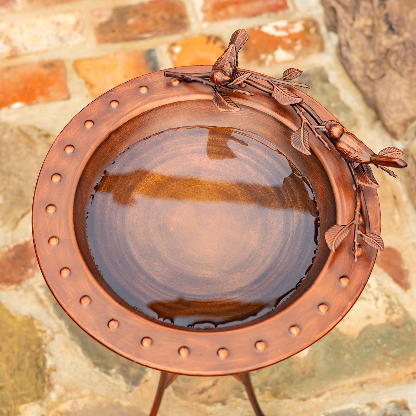 A round copper bird bath with a small metal stand, featuring decorative leaf and bird motifs along the rim, shown filled with water against a rustic stone and brick background.