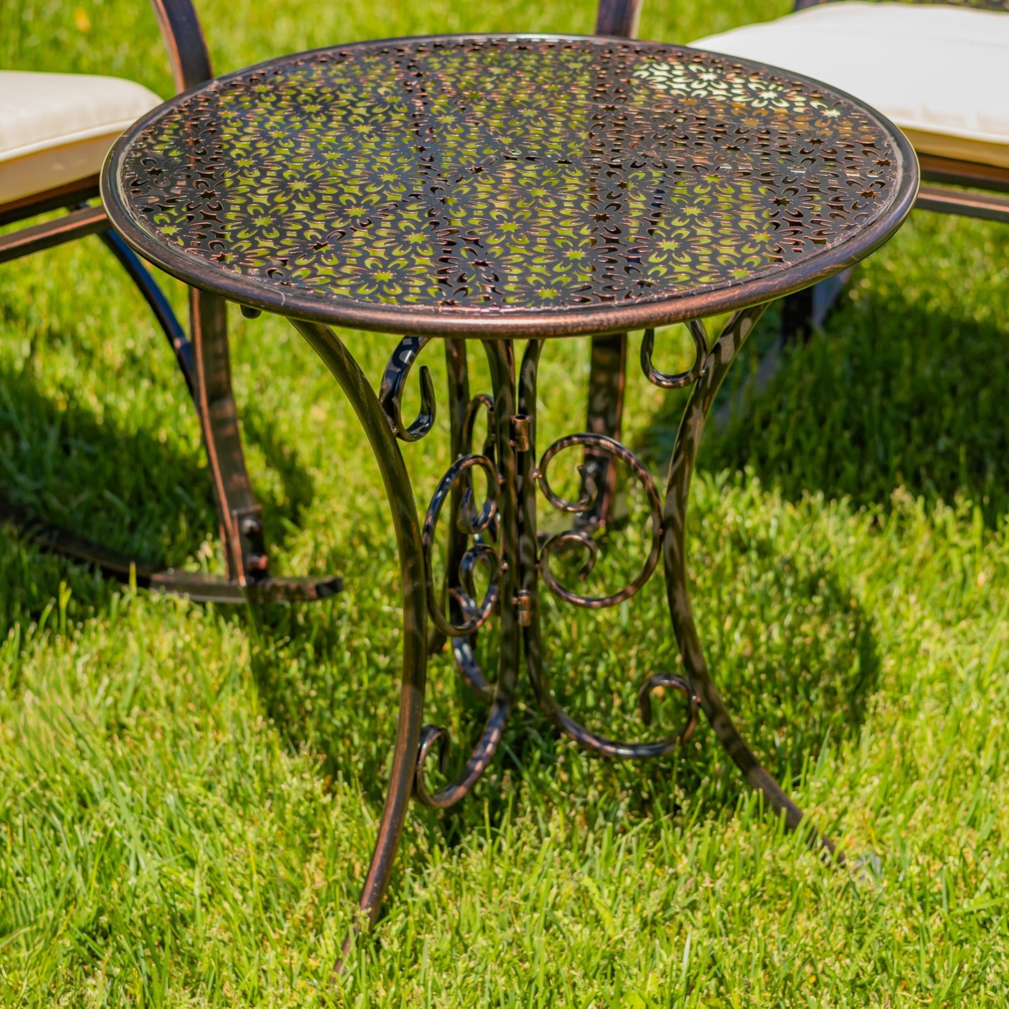 A round metal patio bistro table with a decorative perforated top and ornate scrollwork base, shown on a green lawn outdoors.