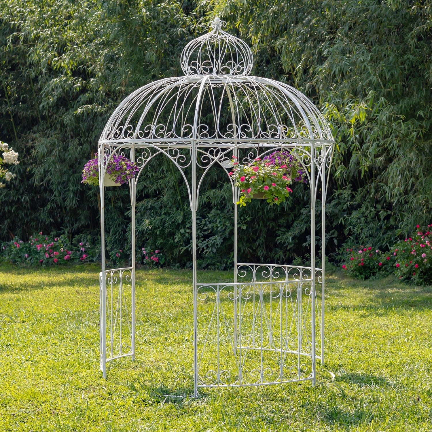 A white wrought-iron garden gazebo with hanging flower baskets, set on a green lawn with a dense green hedge background.