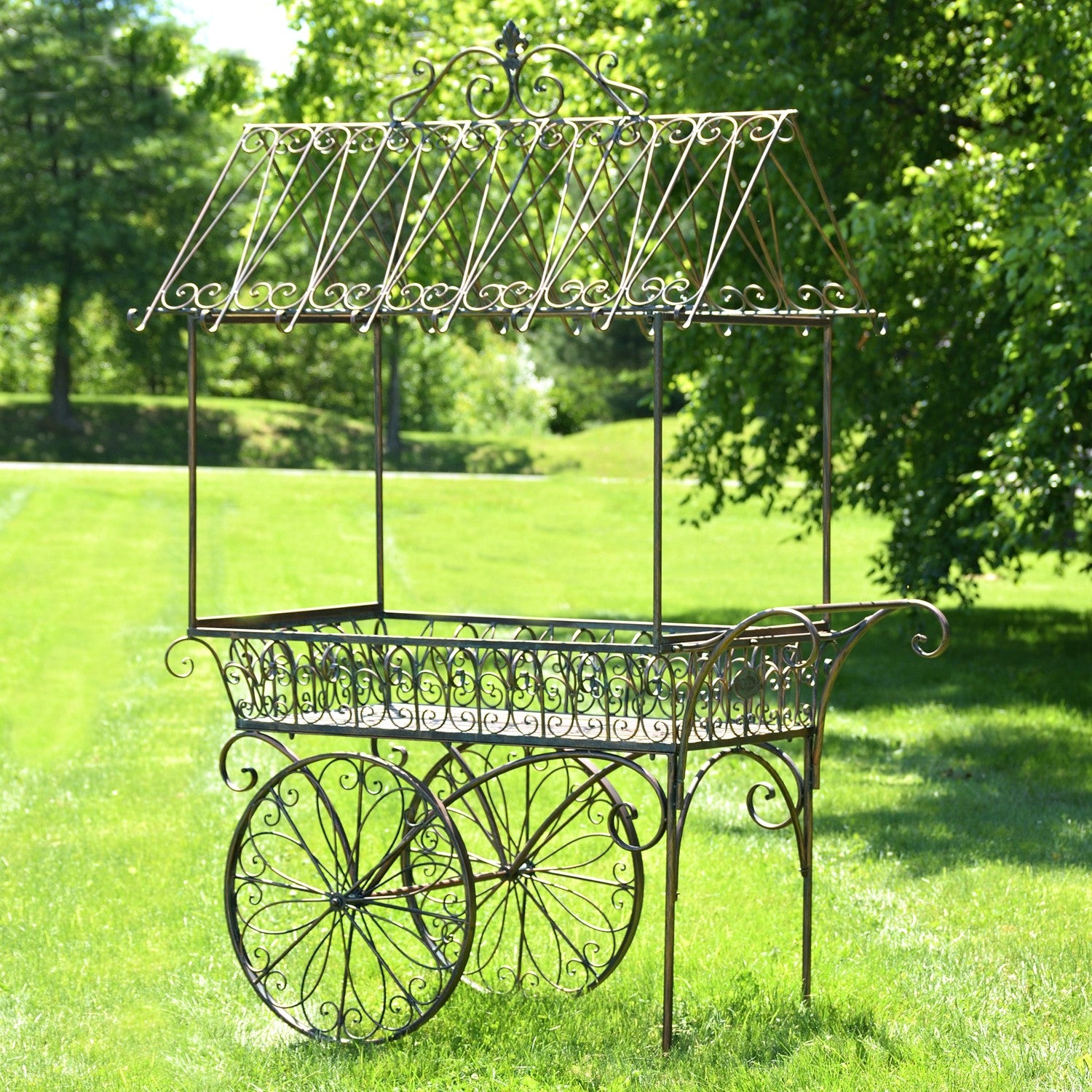 A decorative wrought-iron garden cart with a canopy roof and large spoked wheels, photographed outdoors on a green lawn with trees in the background.