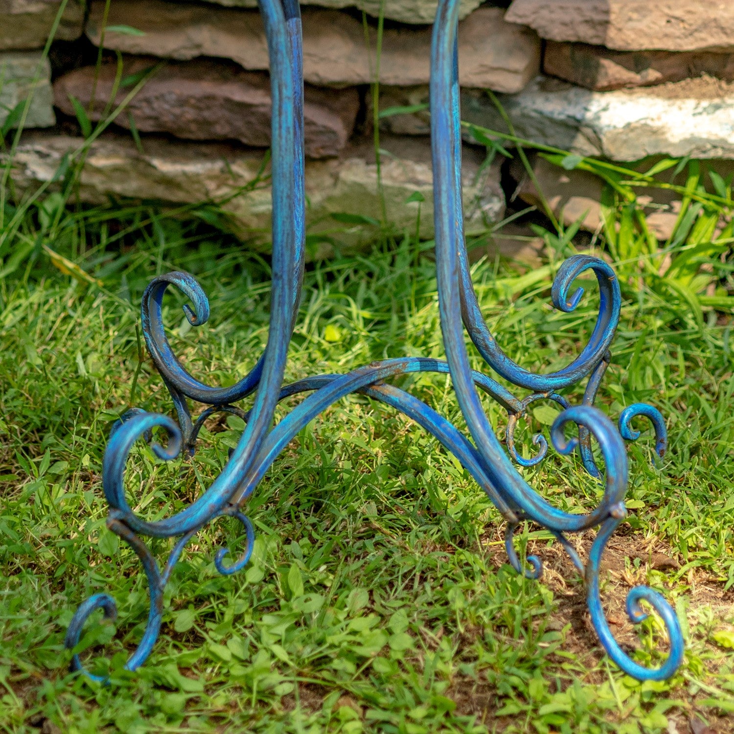 A blue-painted wrought iron garden table base with ornate scrollwork legs, shown standing on green grass in front of a stone wall.