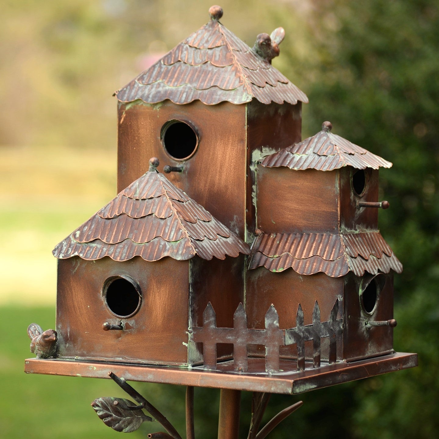 A rustic multi-tiered copper birdhouse with a weathered bronze finish, featuring three stacked levels, decorative birdhouse-roof shapes, small bird figures, and a picket fence detail, set against a blurred green garden background.