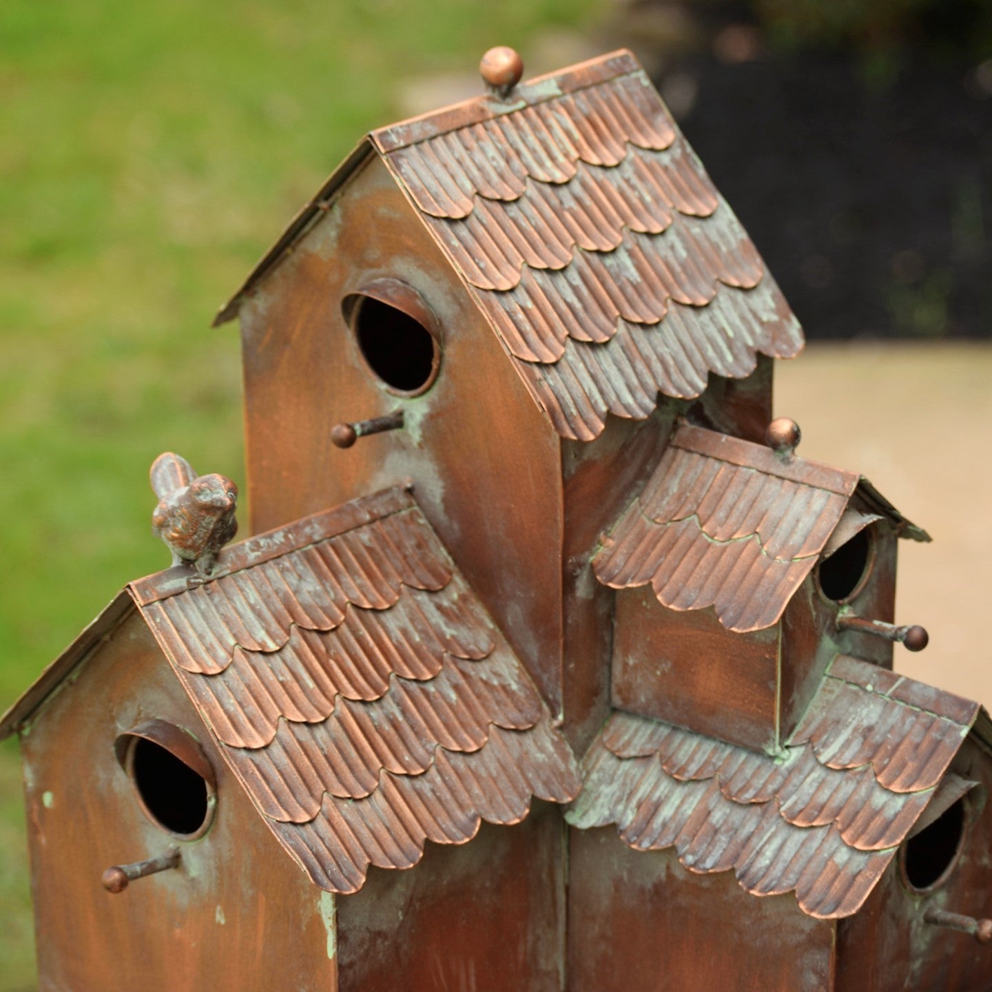 A close-up of a rustic multi-tiered birdhouse structure made of weathered copper-toned metal with scalloped roof tiles and round entry holes, photographed outdoors against a blurred green and beige background.