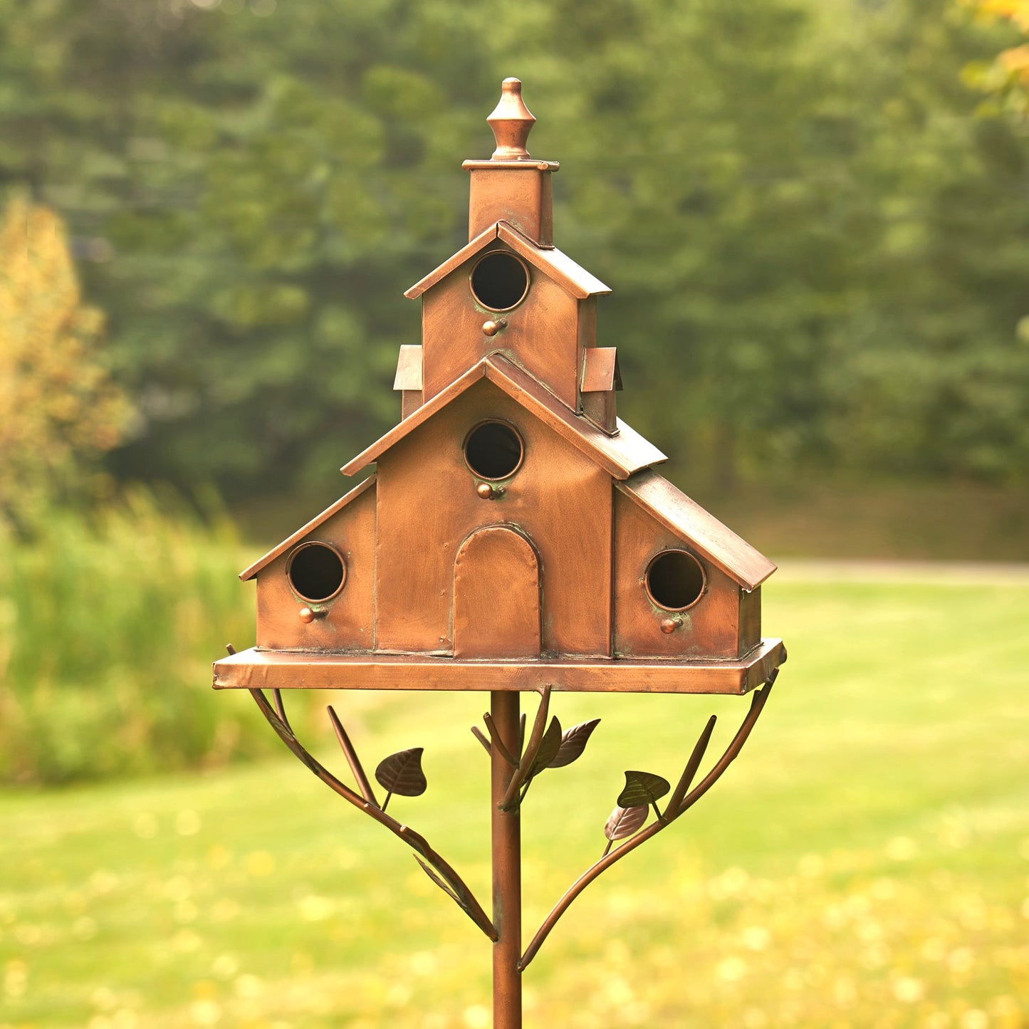 A copper birdhouse mounted on a decorative metal stake with leaf-shaped branches, photographed outdoors against a blurred green garden background.