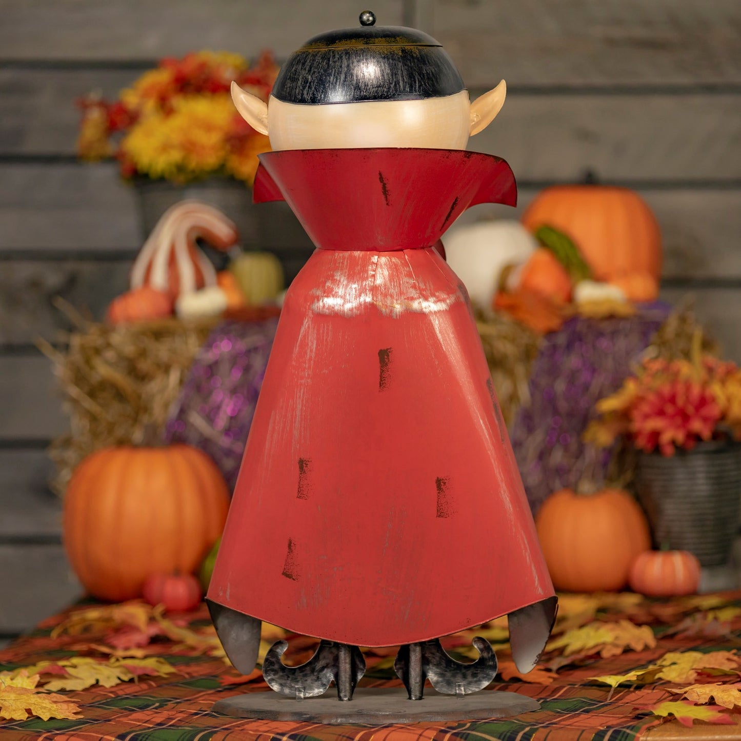 A Halloween decorative vampire figurine shown from the back, wearing a red cape and black hat, standing on a table with pumpkins, hay bales, and autumn flowers in the background.