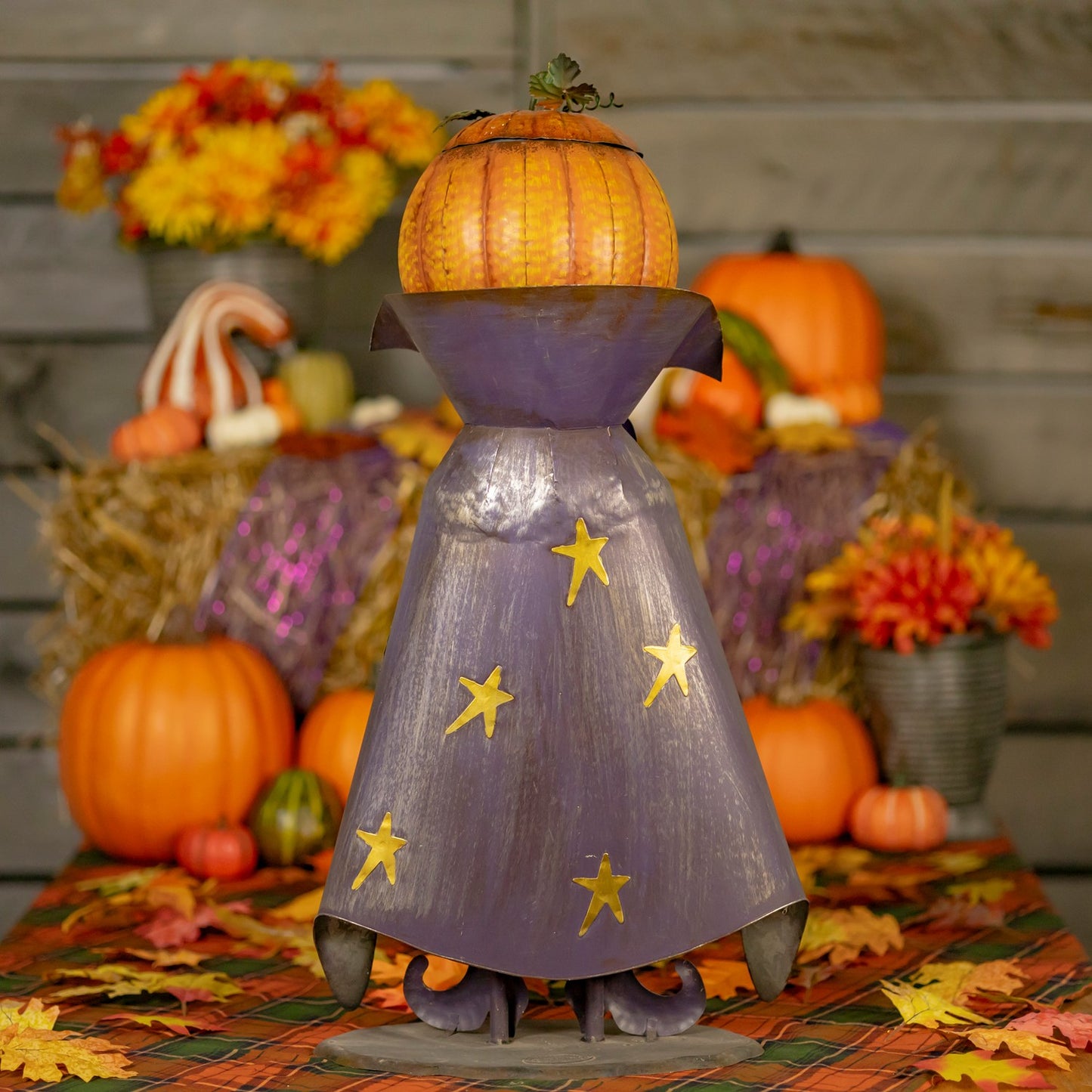 A decorative autumn metal figurine shaped like a wizard's hat or cloaked figure, with a pumpkin on top, a purple body, and gold stars, displayed on a table with pumpkins, fall leaves, and floral arrangements in the background.