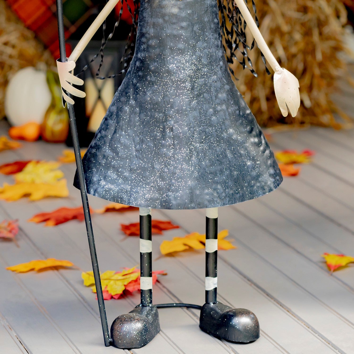 A Halloween decorative figure with a glittery dark blue dress, striped black-and-white legs, and a cane, shown standing on a gray plank floor with scattered autumn leaves and fall decor in the background.
