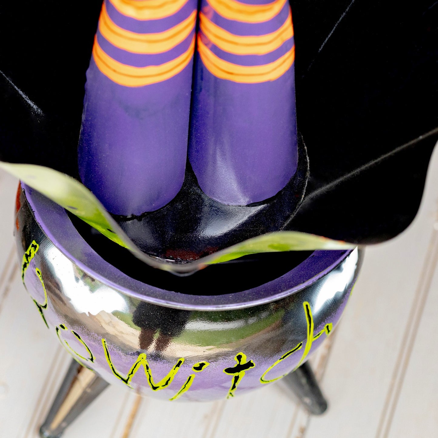 A close-up of a witch-themed product, likely a decorative planter or bowl, with a purple interior, black exterior, green script lettering, and a pair of purple socks with orange stripes resting inside.