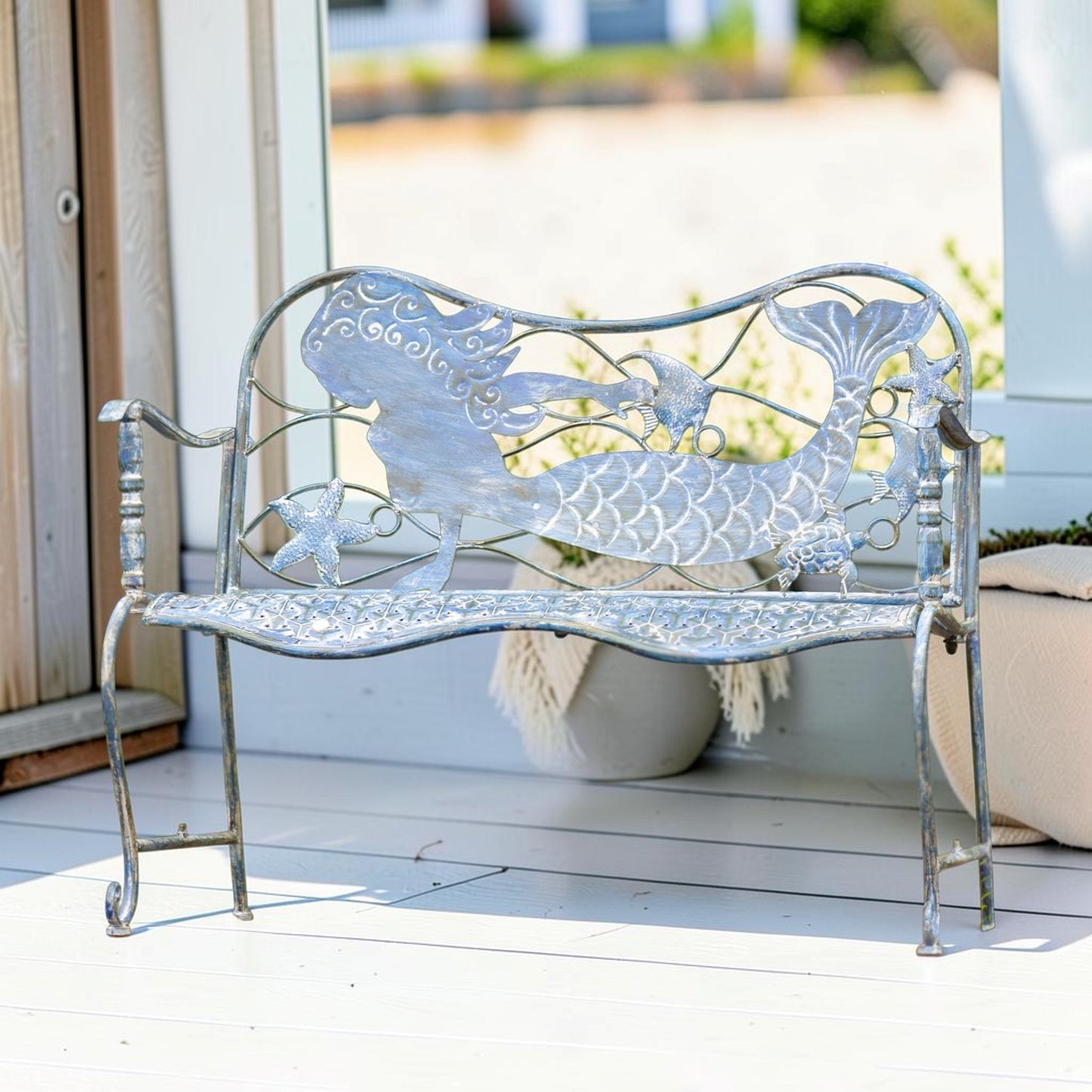 A decorative metal garden bench with a mermaid and starfish design, painted in a distressed silver finish, set on a white porch.