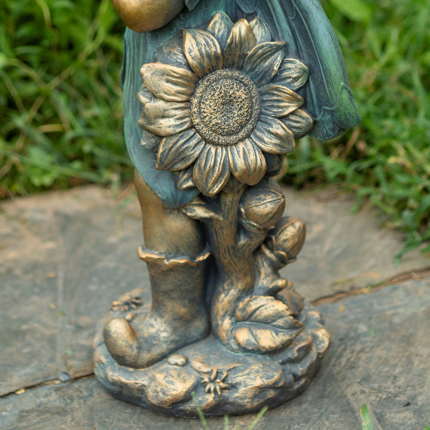 Close-up of a bronze-toned garden statue showing a sunflower, buds, leaves, and part of a child's legs and dress, placed outdoors on a stone surface with grass in the background.
