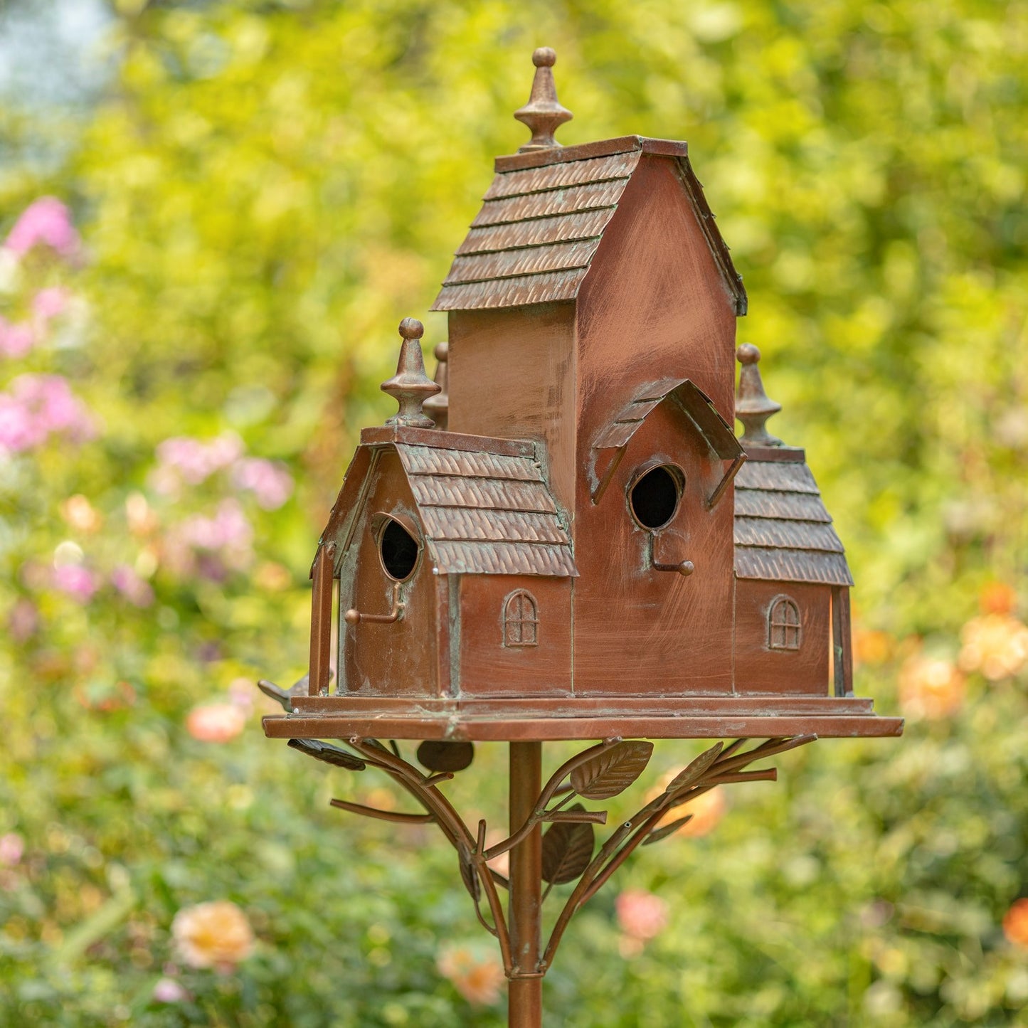 A decorative multi-tiered birdhouse with a weathered copper or bronze finish, mounted on a matching metal stand with leaf details, set against a blurred green garden background.