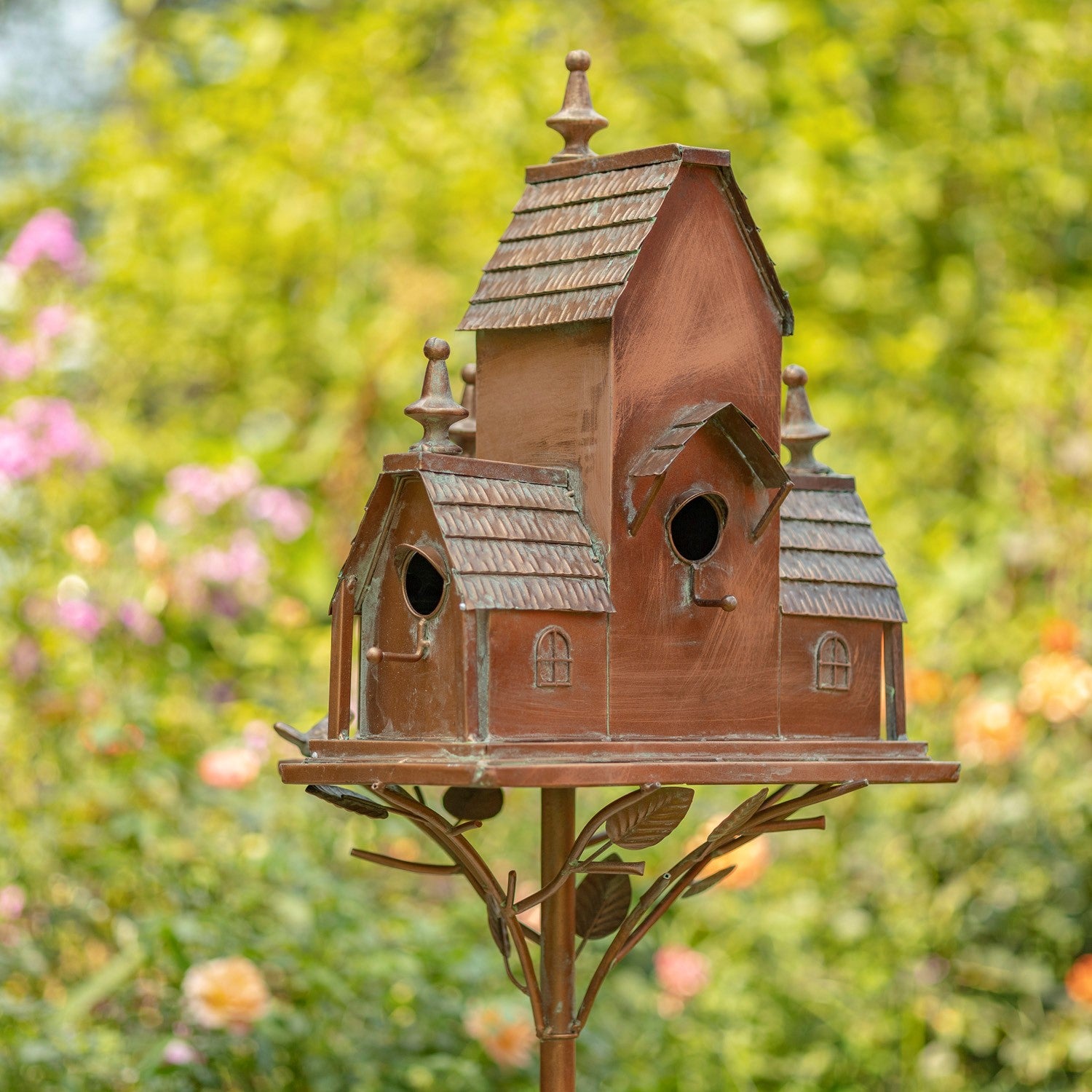 A decorative multi-tiered birdhouse with a weathered copper or bronze finish, mounted on a matching metal stand with leaf details, set against a blurred green garden background.