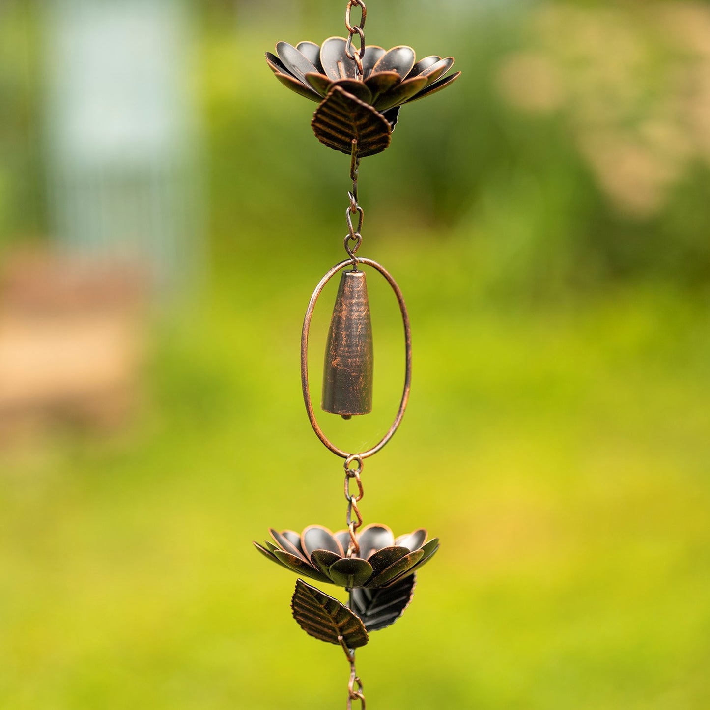 A hanging metal wind spinner garden ornament with two lotus flower shapes and a central hanging cone, photographed outdoors against a blurred green garden background.