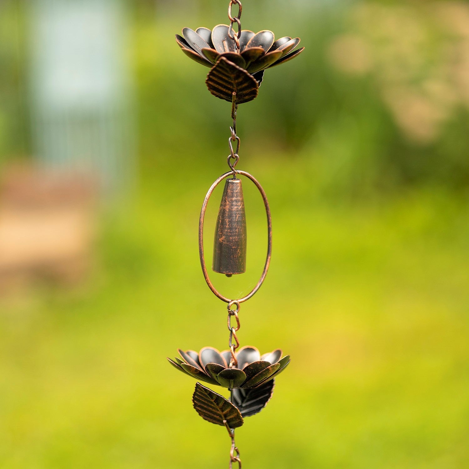 A hanging metal wind spinner garden ornament with two lotus flower shapes and a central hanging cone, photographed outdoors against a blurred green garden background.