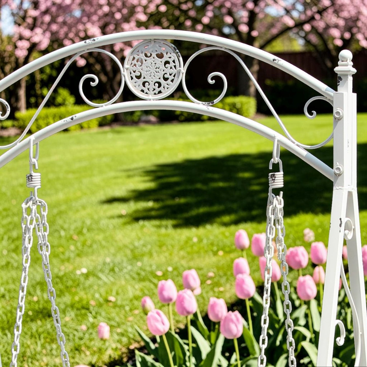 A white wrought iron garden swing with hanging chains, photographed outdoors on a green lawn with pink tulips and pink blossoms in the background.