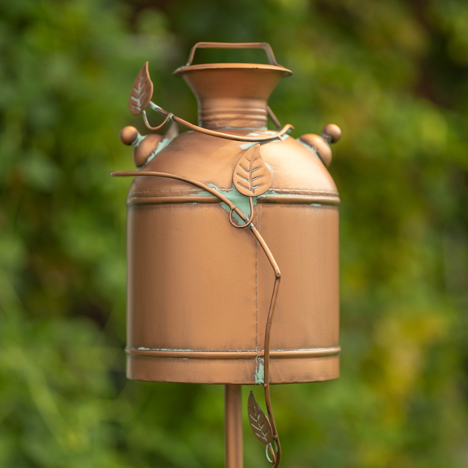 A copper-toned metal garden stake or decorative post shaped like a vintage milk canister, with leaf-shaped accents and a blurred green garden background.
