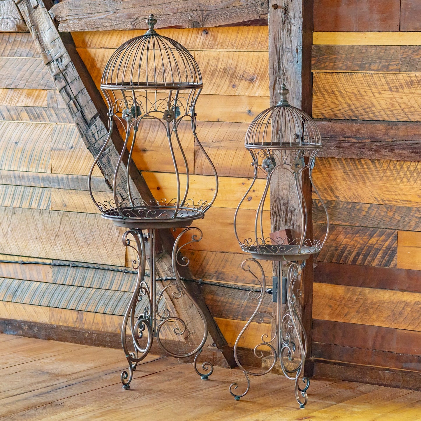 A rustic metal birdcage-style candle holder with a small pedestal table and two hanging candle shades, shown against a wooden plank wall and floor.