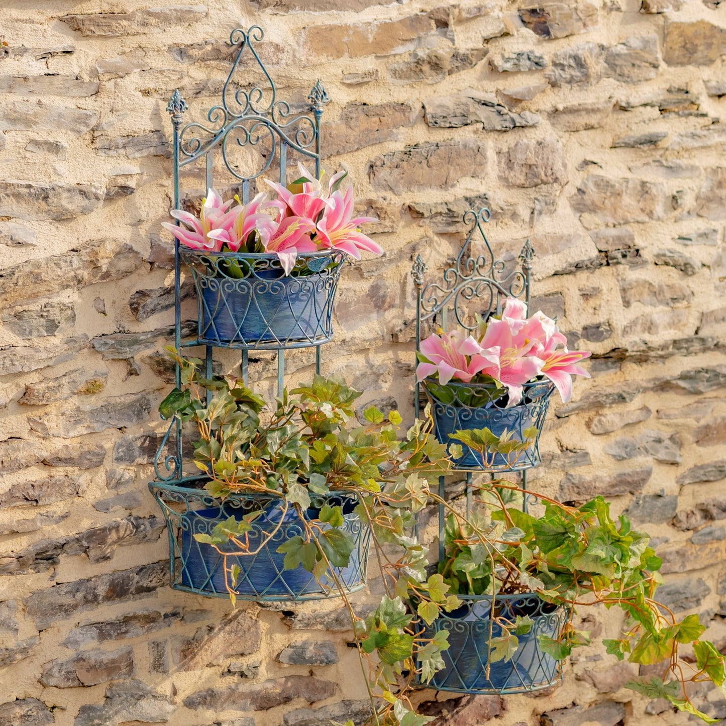 Two blue metal tiered wall planters with ornate scrollwork, mounted on a rustic stone wall. Each planter holds pink lilies and trailing green foliage.