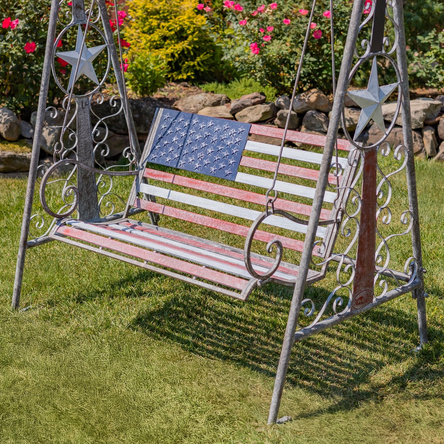 A metal swing bench with an American flag design, set on a green lawn in a garden. The swing features a distressed red, white, and blue striped seat and backrest, with a blue star-spangled panel on the left side.