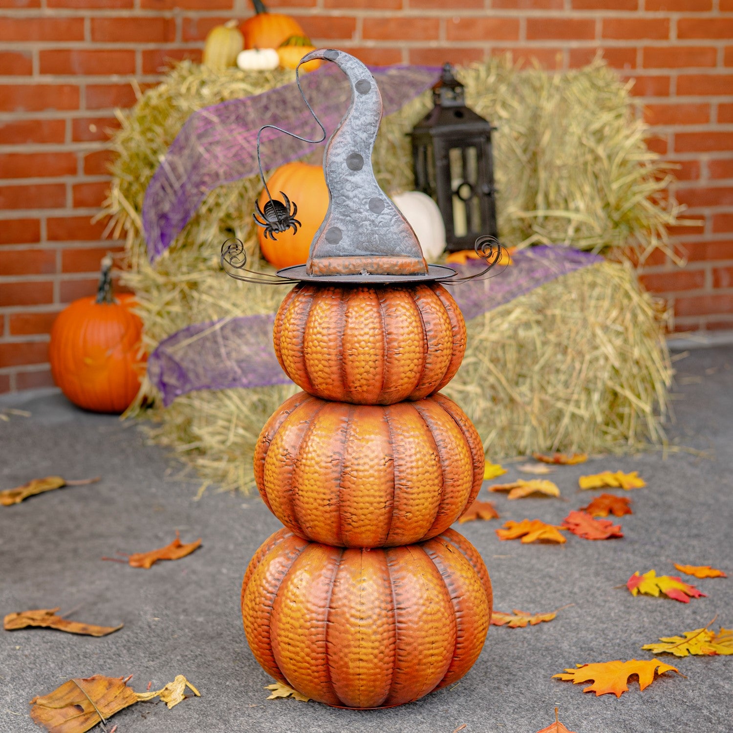 Decorative pumpkin stack with a witch hat and small pumpkin on top, set against a brick wall with hay bales and pumpkins.