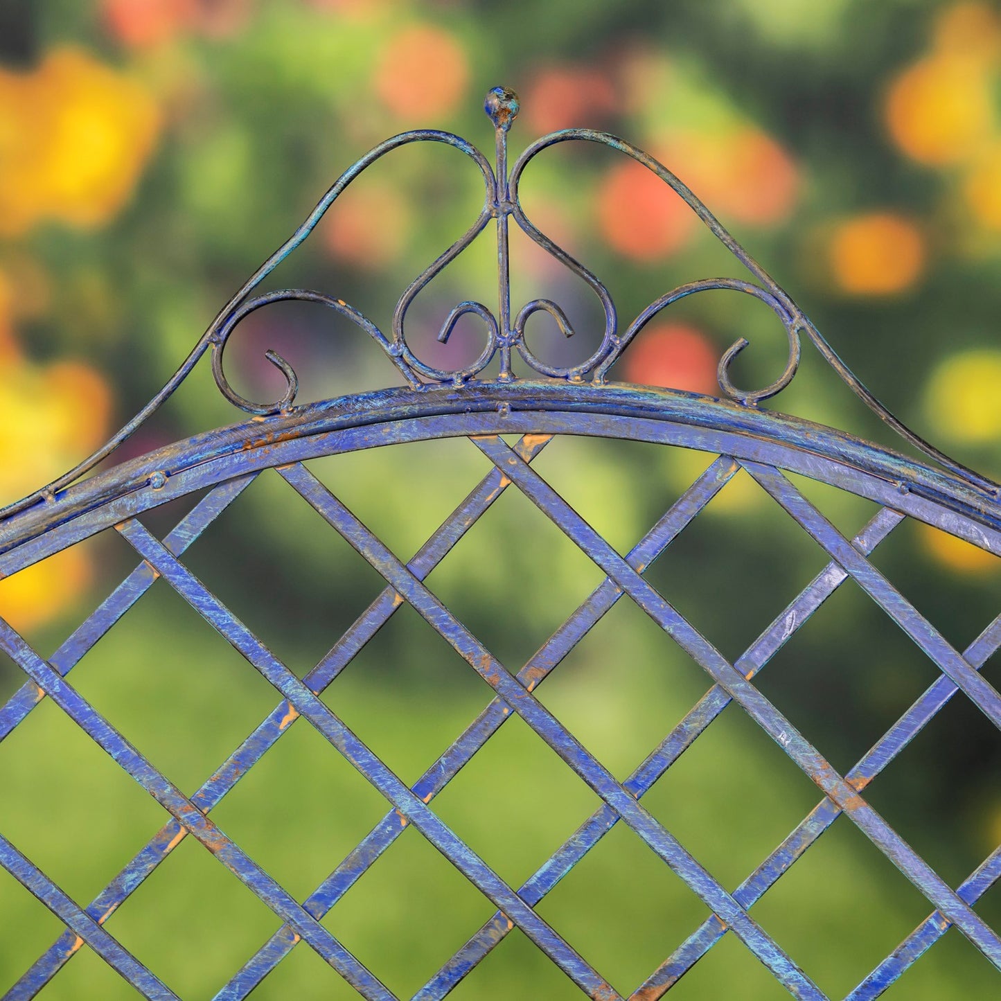 Close-up of an ornate blue metal garden gate with a diamond lattice pattern and decorative scrollwork at the top, set against a blurred green and yellow outdoor background.