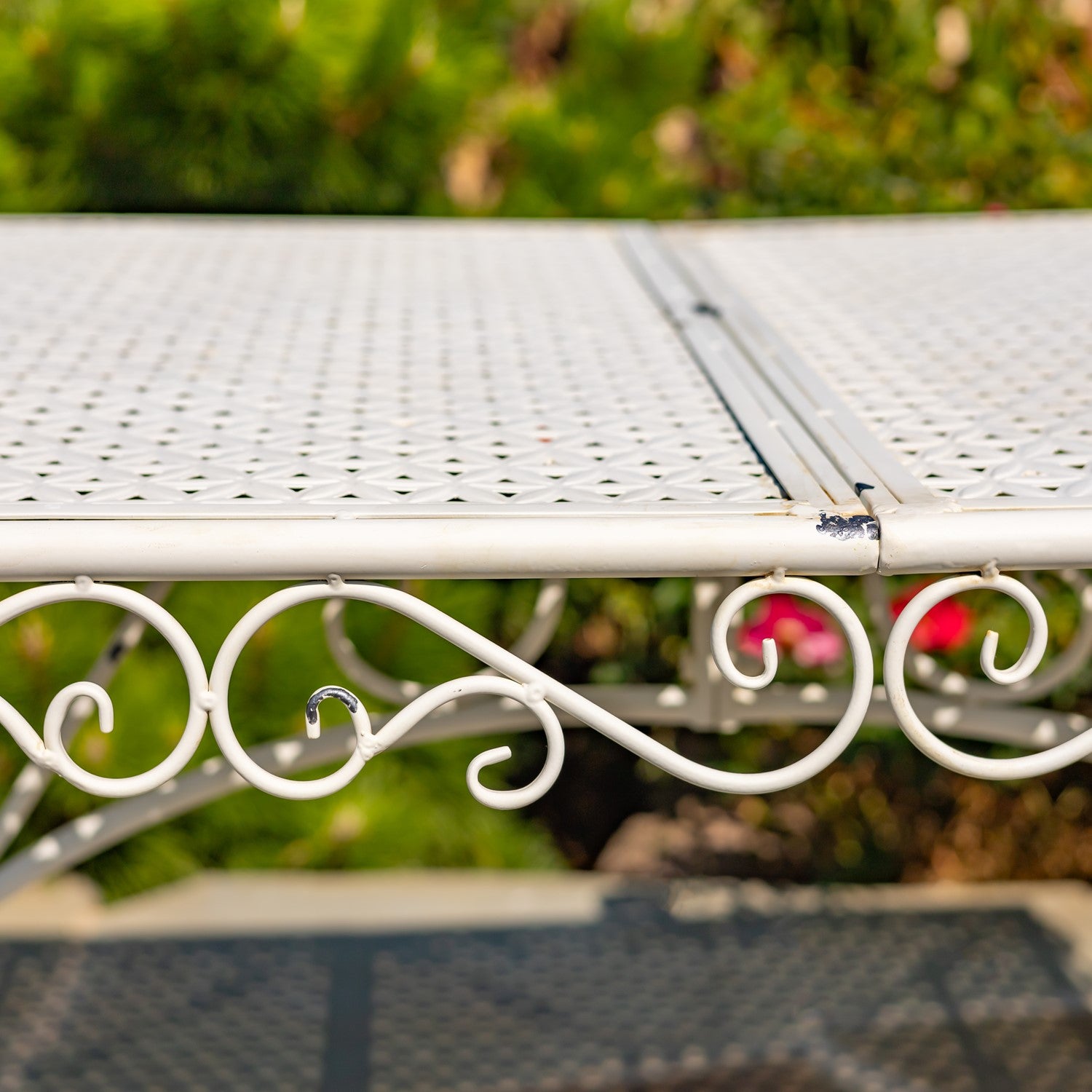 White metal patio table with a perforated slatted top and decorative scrollwork base, photographed outdoors with blurred greenery in the background.
