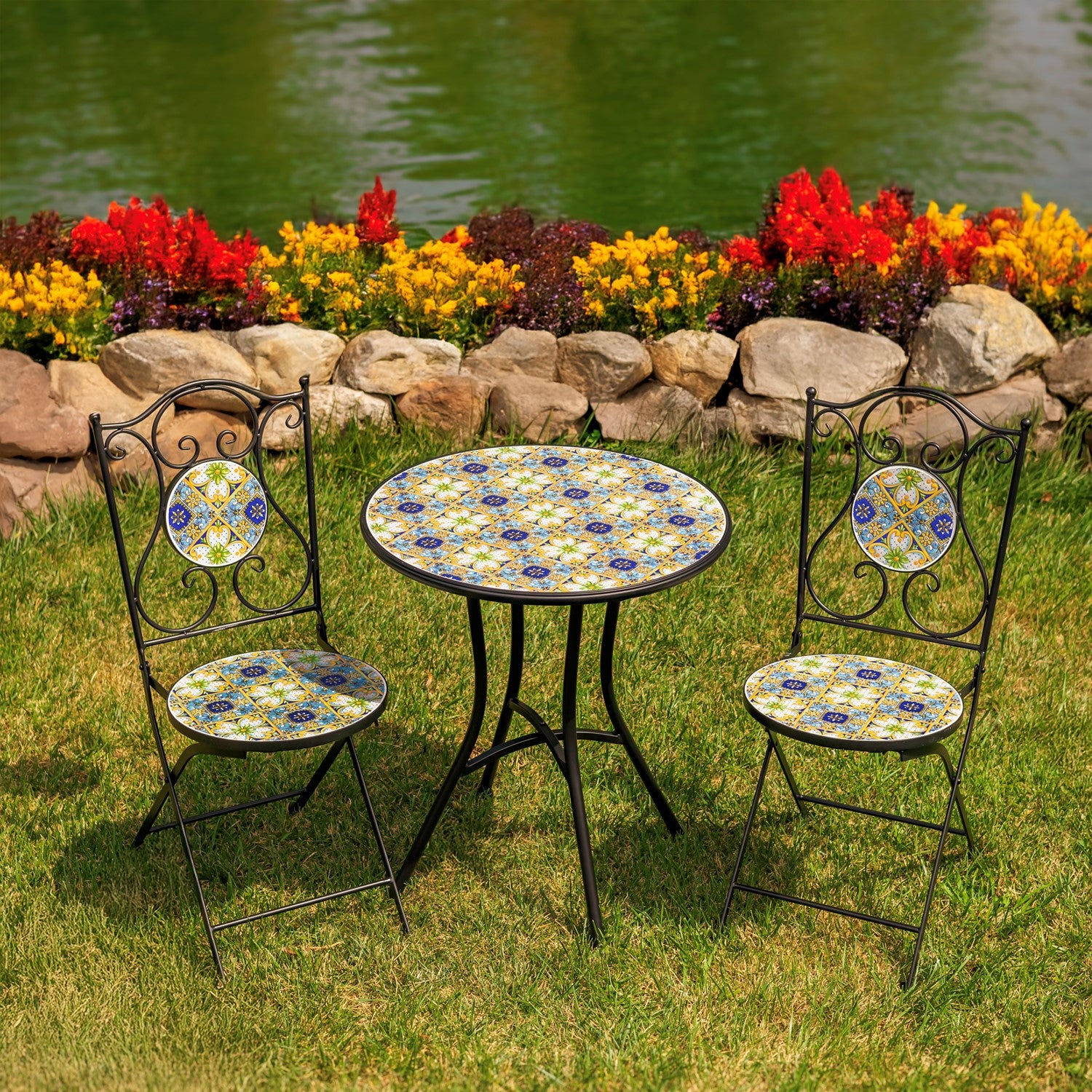 A 3-piece outdoor patio set featuring a round metal table and two matching chairs with decorative blue, yellow, and white mosaic tile backs and seats, set on a green lawn in front of a flower bed and lake.
