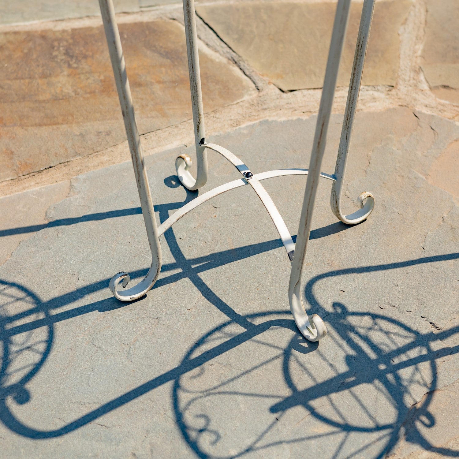 A silver metal plant stand with a decorative scrollwork base, photographed outdoors on a stone patio with sharp shadows cast across the surface.