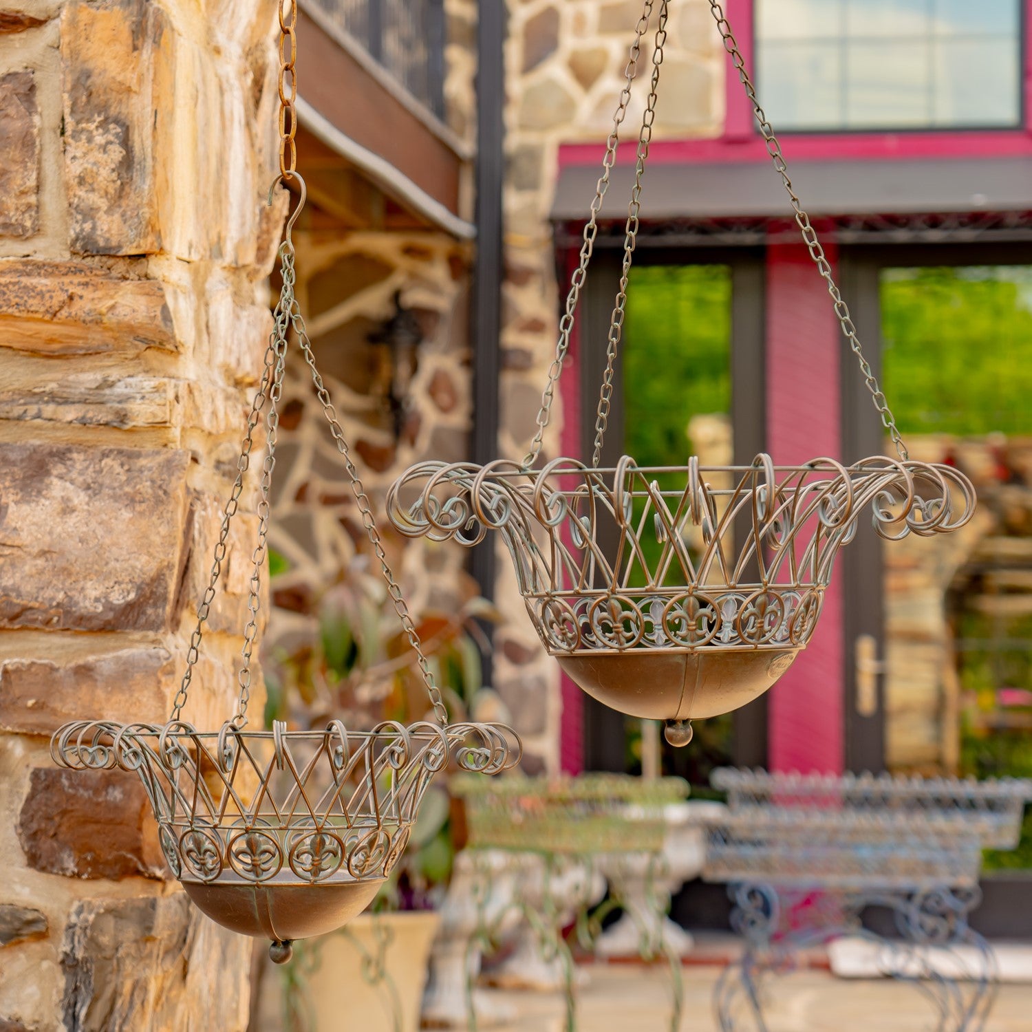 Two ornate hanging metal planters with scrollwork tops and bowl-shaped bases, suspended by chains outdoors against a stone wall and blurred garden background.