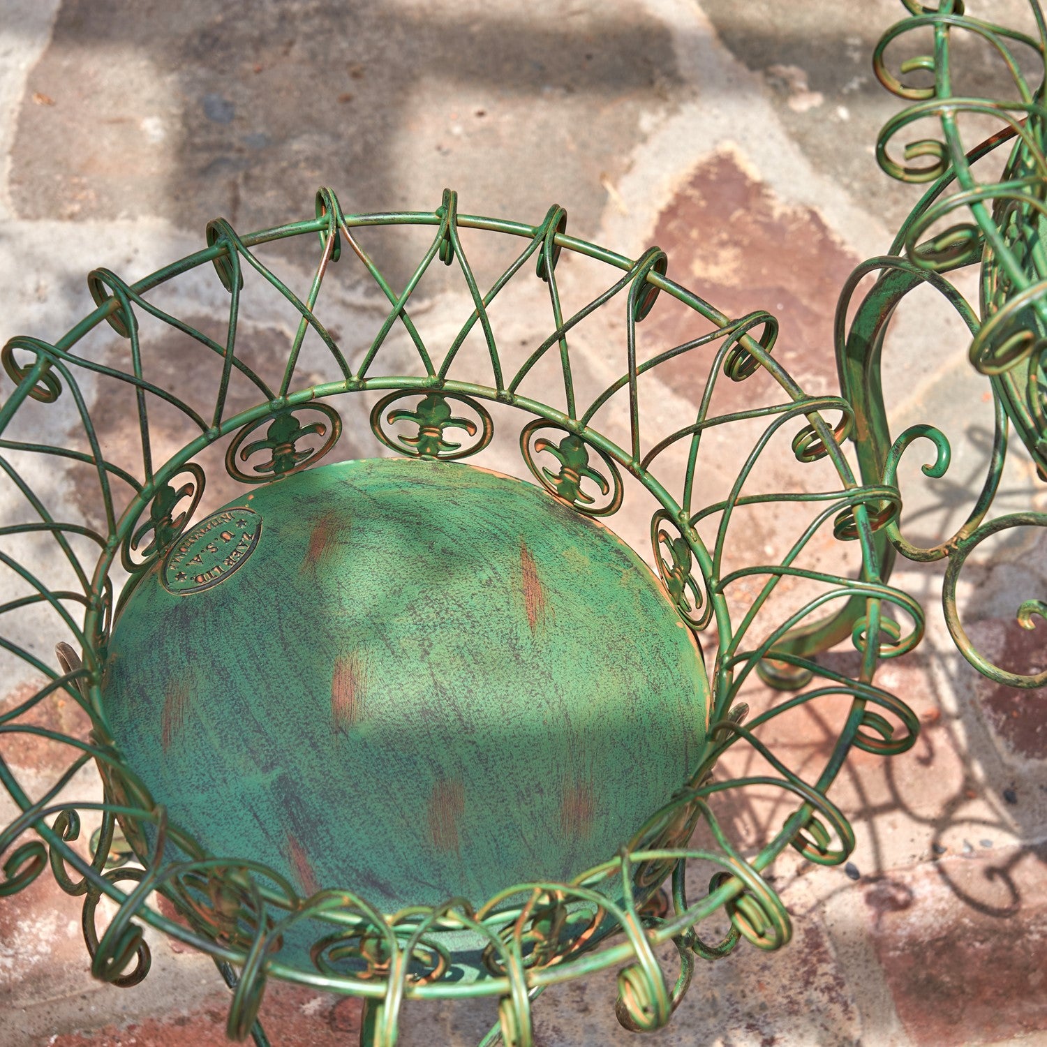 A green metal garden basket with an intricate scrollwork design and a distressed teal-green patina inside, photographed outdoors on a stone paver surface.