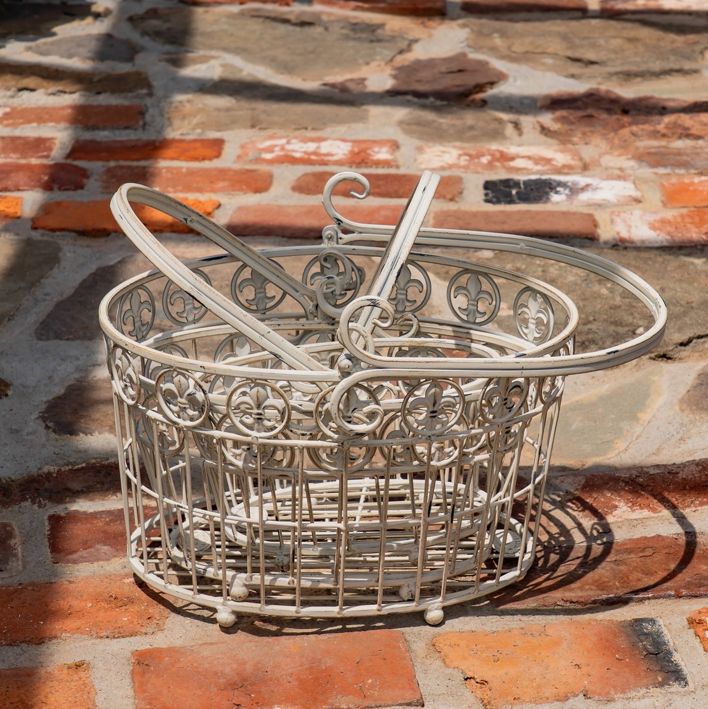 A white-painted metal wire basket with decorative scrollwork and a hanging loop handle, placed on a rustic red brick and stone surface.
