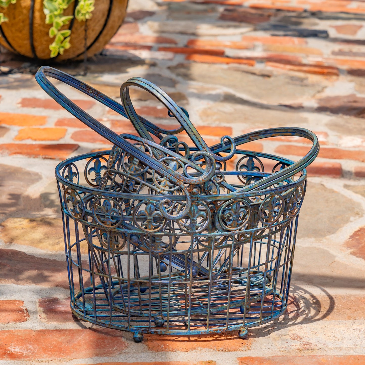 A decorative metal wire basket with ornate scrollwork and looped handles, shown outdoors on a stone patio. The basket has a distressed blue-green patina with brown undertones.
