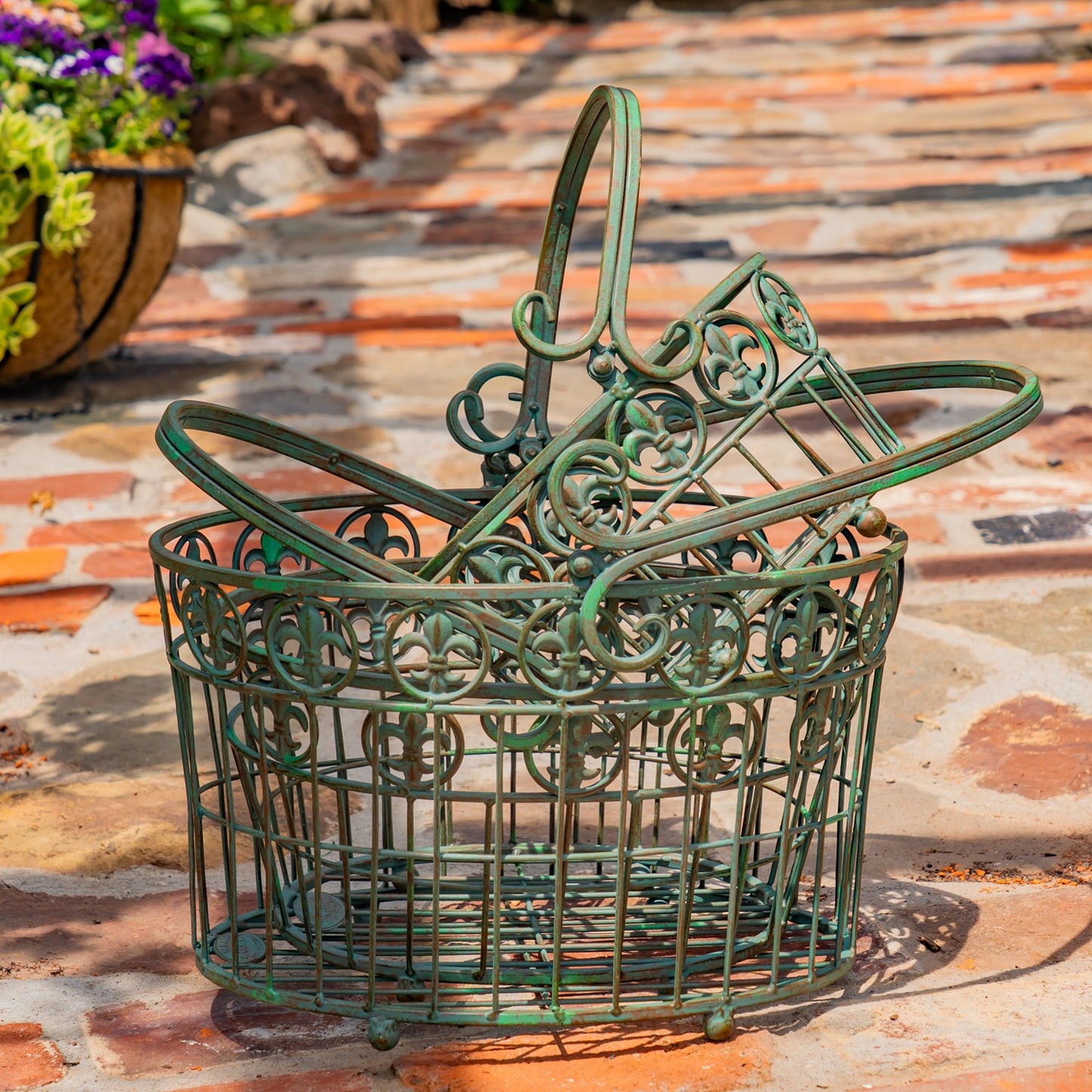 A green metal wire garden basket with an ornate scrollwork design and a tall handle, placed on a stone patio outdoors.