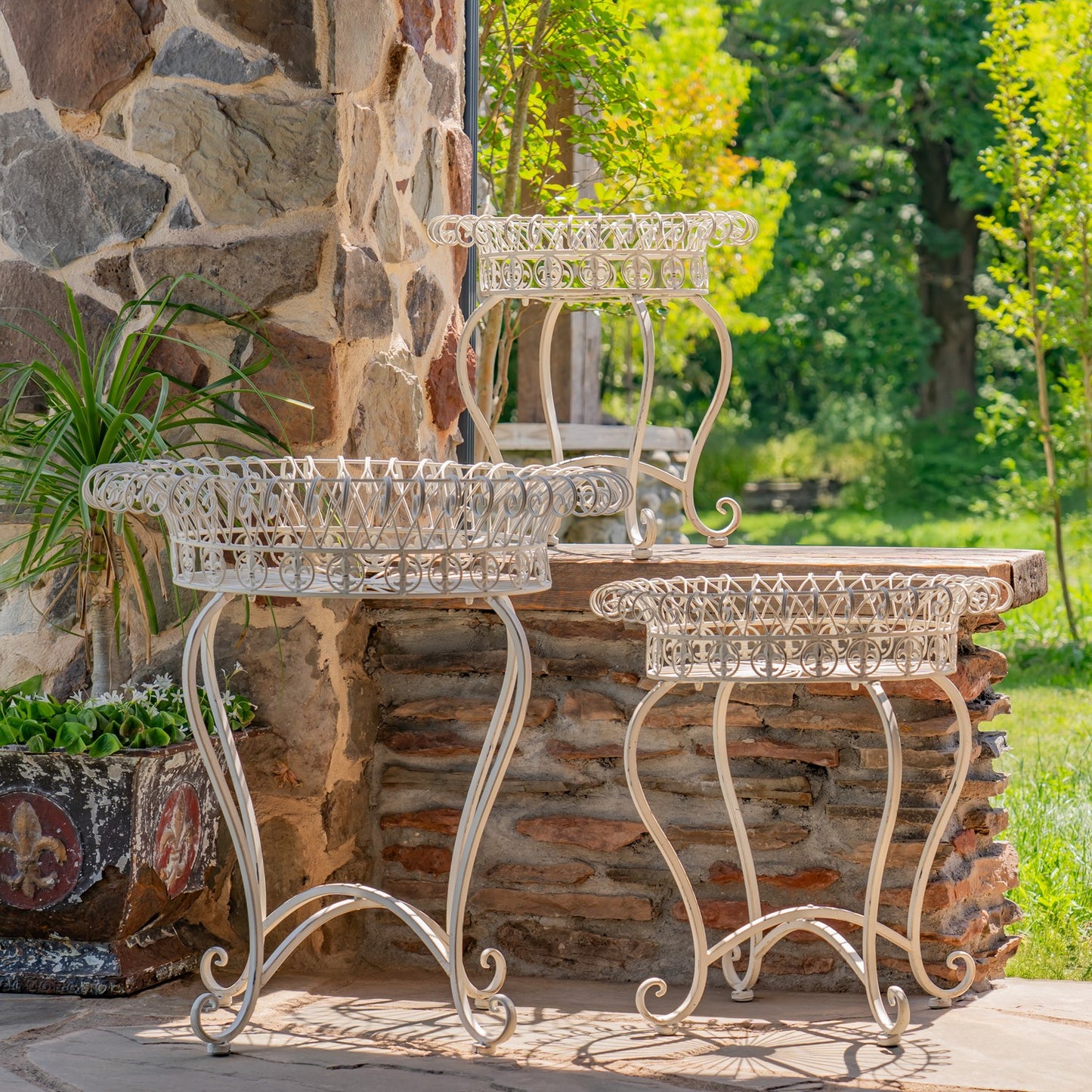 Three white metal garden planters in different sizes arranged on a stone patio in front of a rustic stone wall with green grass and trees in the background.