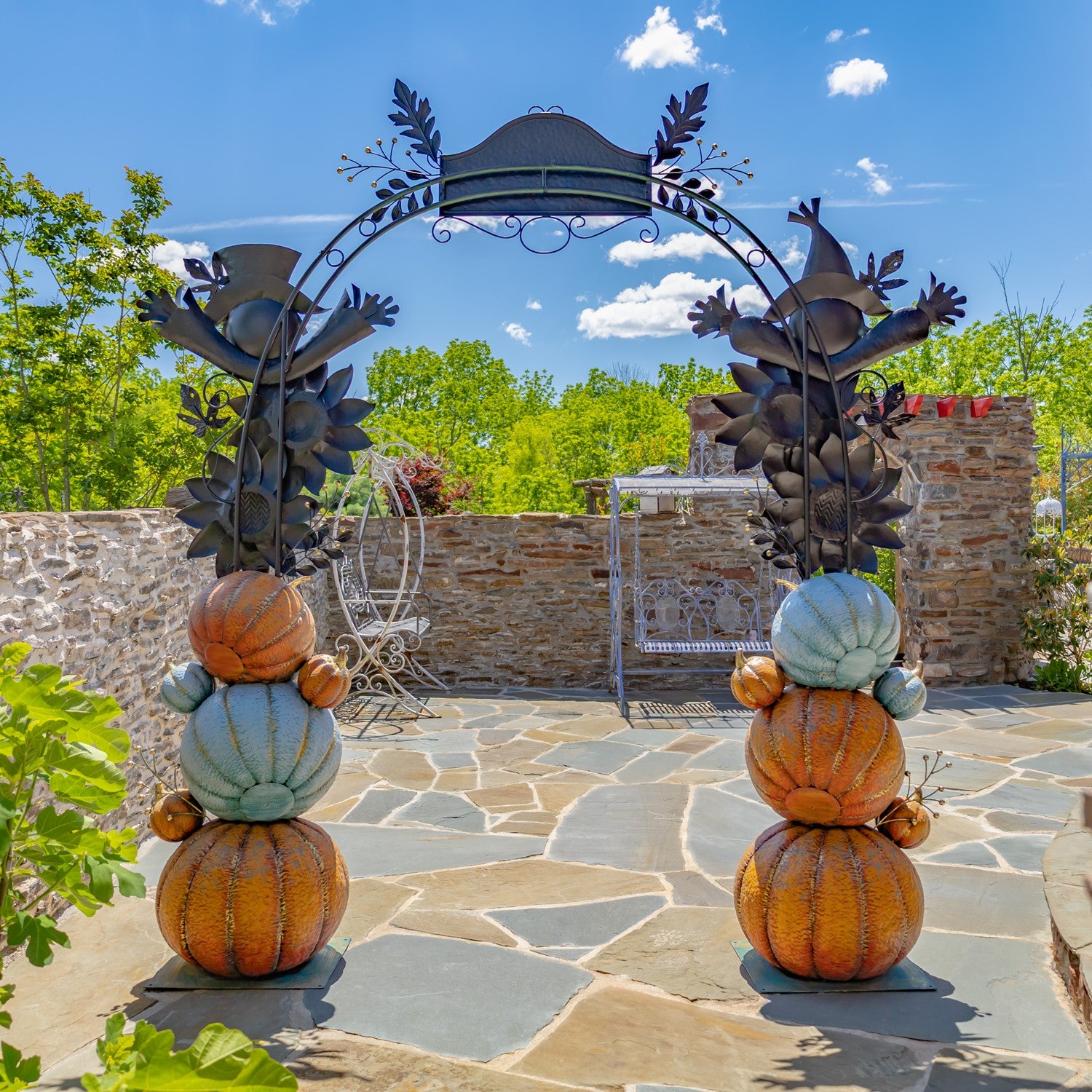 Outdoor autumn garden arch decoration with stacked faux pumpkins and gourds in orange, tan, and pale blue, flanked by dark metal leafy gate supports on a stone patio under a blue sky.