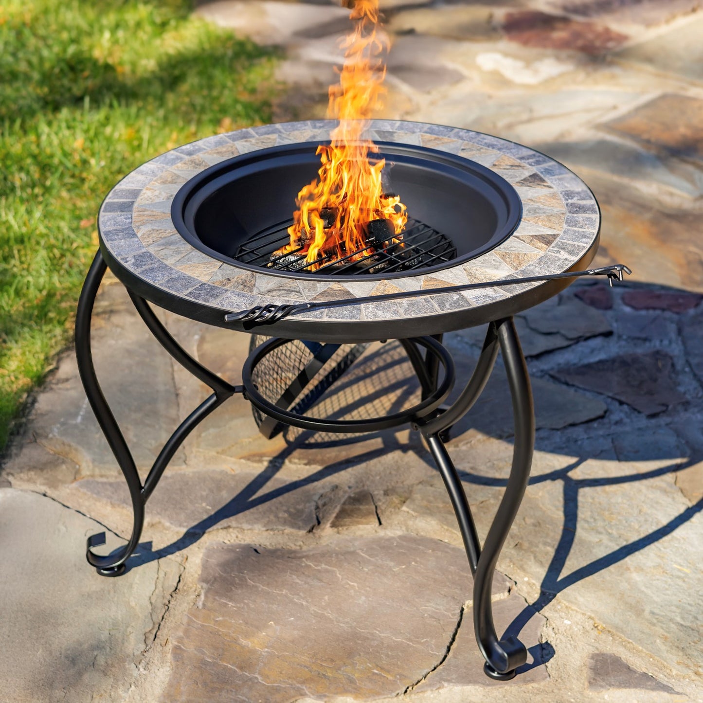 Outdoor fire pit table with a circular stone-tile surround and black metal legs, shown with a visible flame burning in the center on a stone patio.