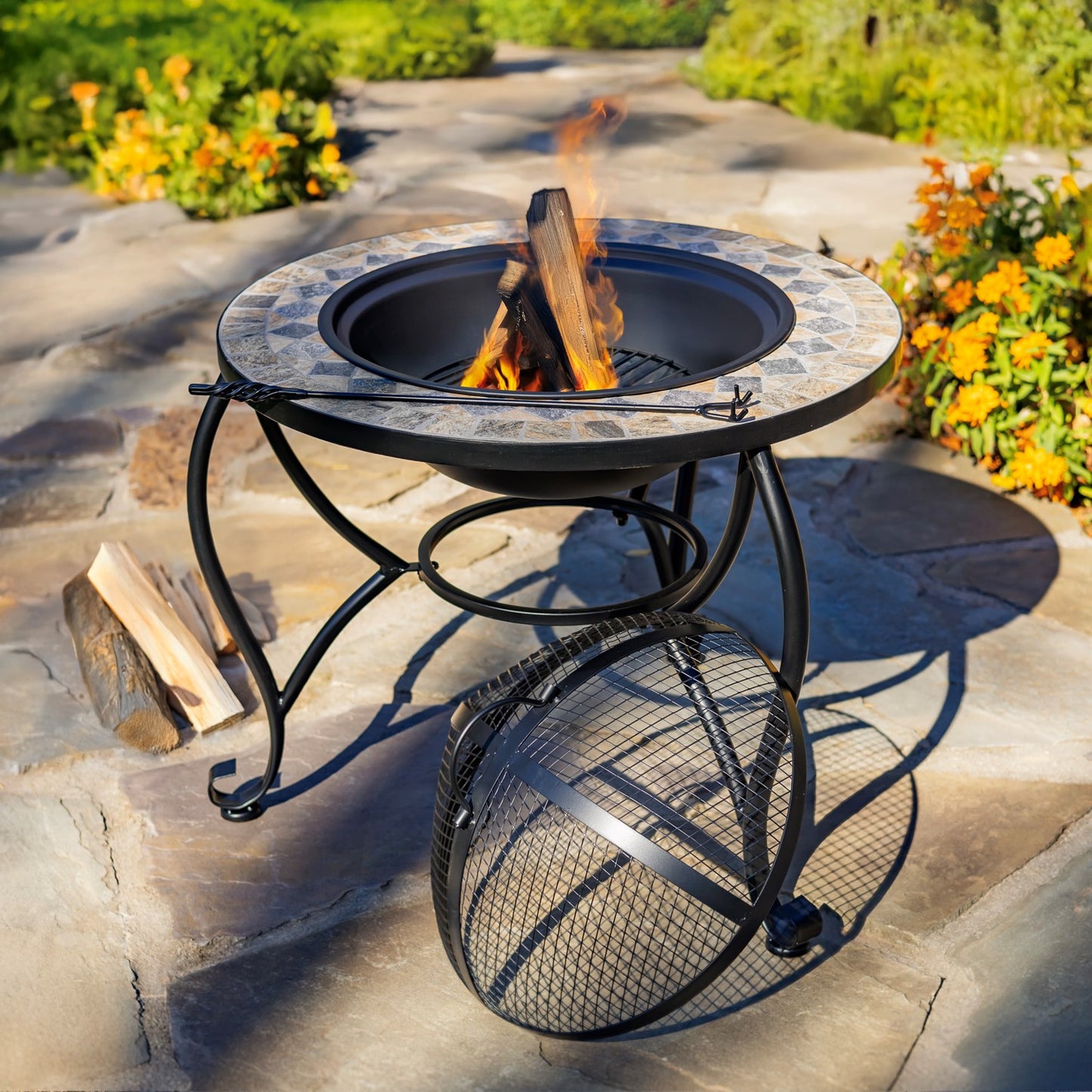 Outdoor fire pit table with a stone-patterned top, black metal base, and hanging mesh screen, shown burning wood on a stone patio with flowers in the background.