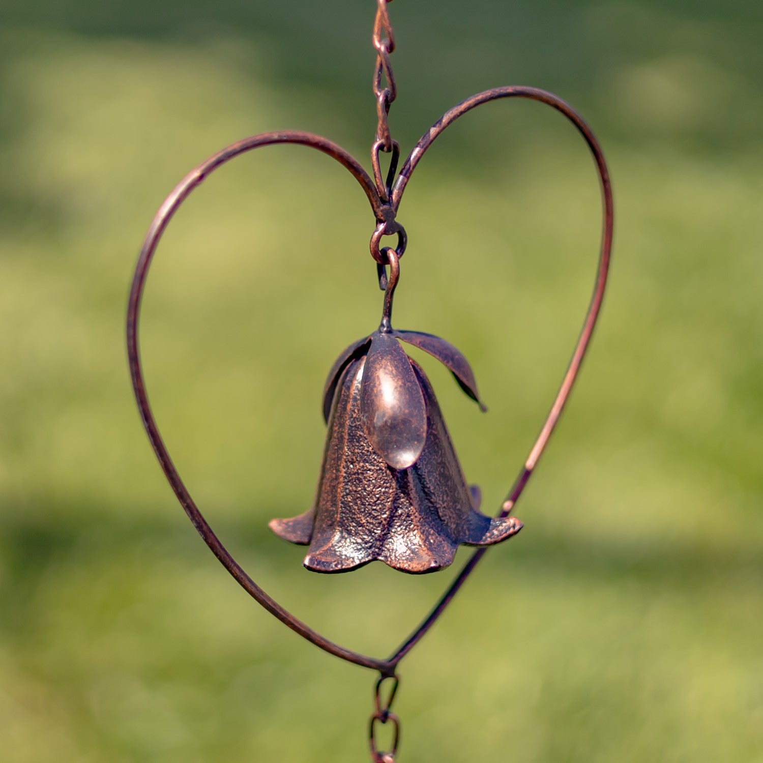 A copper-toned metal garden wind spinner featuring a bell-shaped flower suspended inside a heart-shaped wire frame, set against a blurred green grass background.