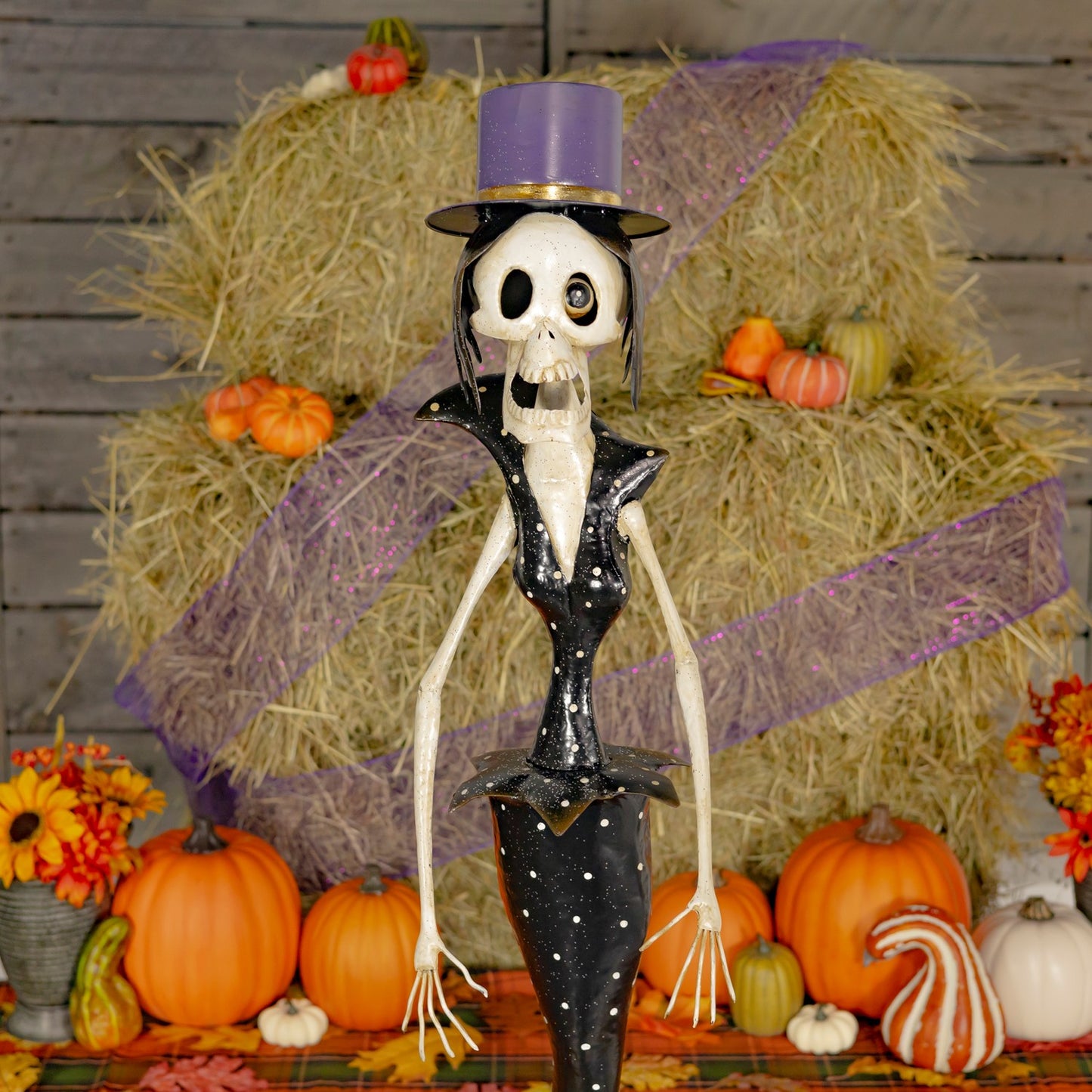 Skeleton figure in formal attire with a purple top hat surrounded by pumpkins and hay bales.