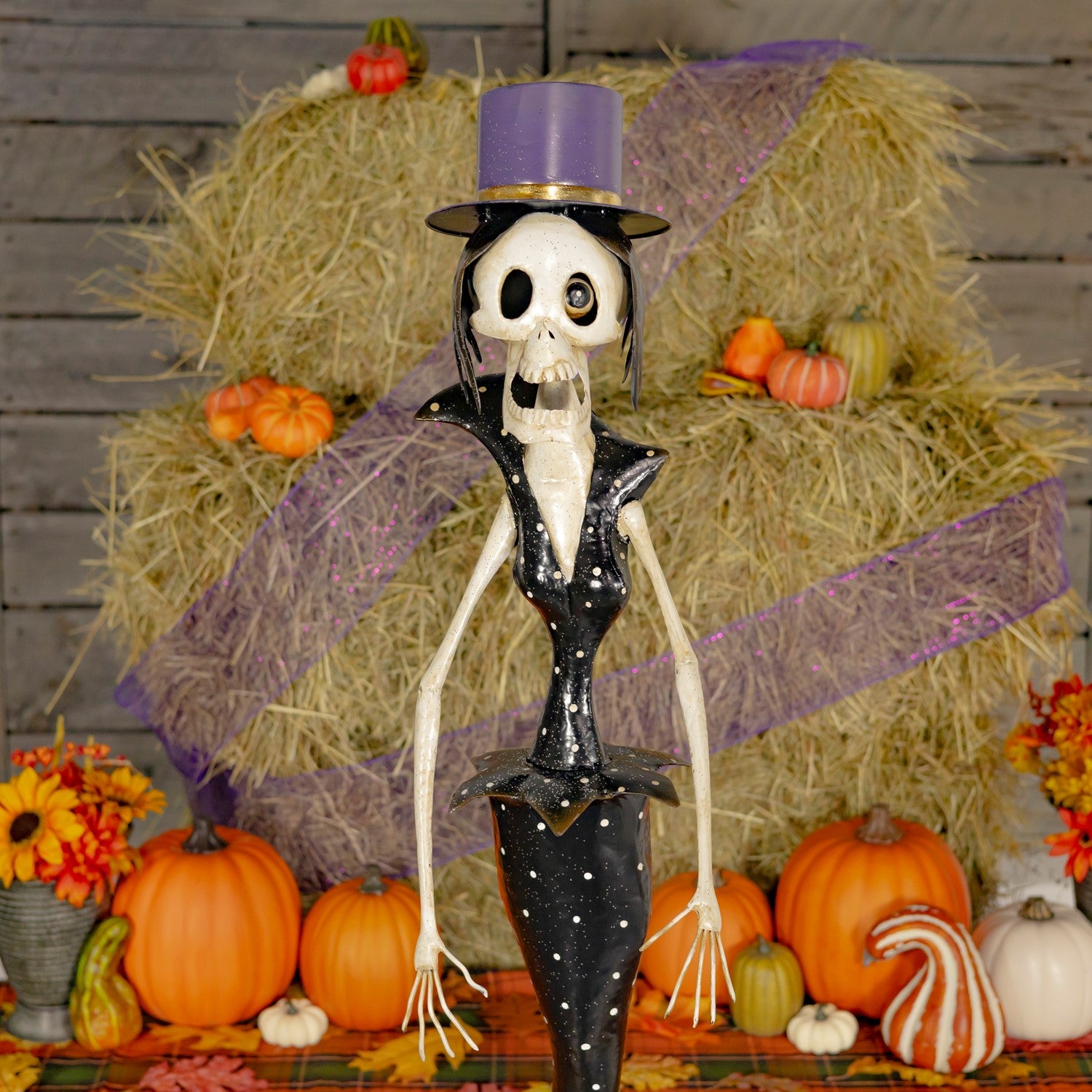 Skeleton figure in formal attire with a purple top hat surrounded by pumpkins and hay bales.