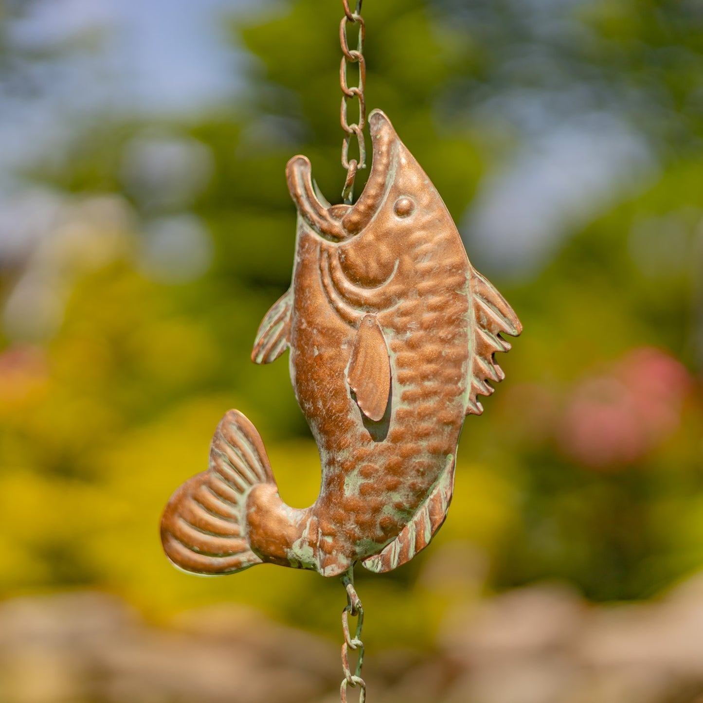 A copper-colored metal fish garden wind spinner hanging from a chain outdoors, with a blurred green and yellow garden background.