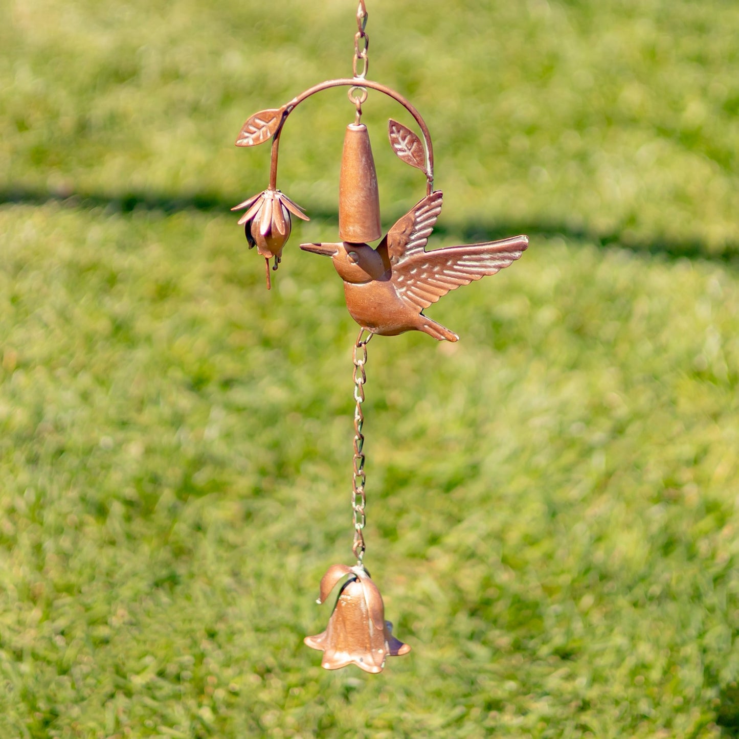 A copper bird wind spinner with a bell and flower details, hanging outdoors against a blurred green grass background.